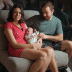 Jeune couple assis sur un canapé gris tenant leur bébé nouveau-né dans un intérieur moderne