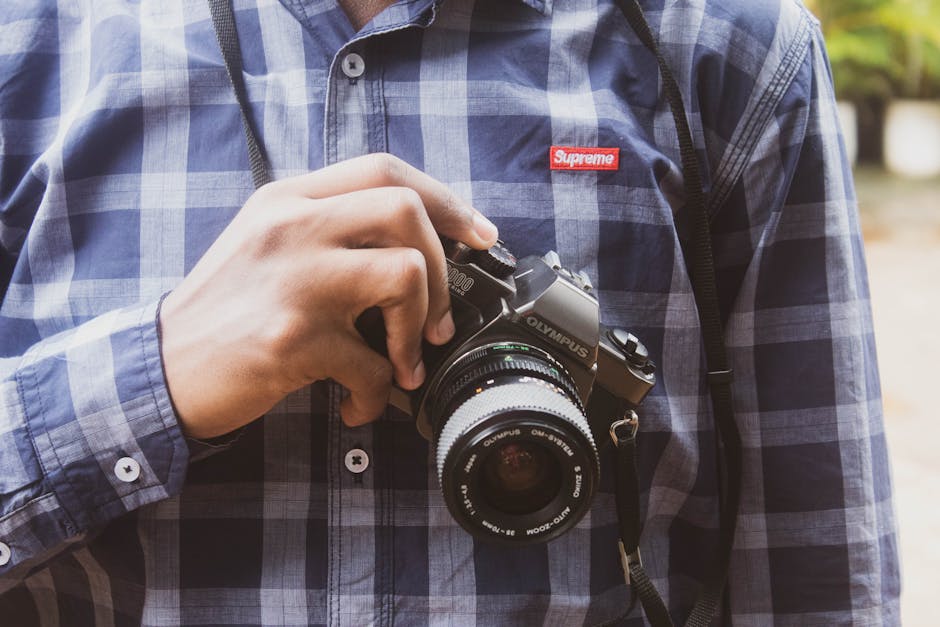 Close-up of a photographer holding an Olympus camera wearing a plaid shirt outdoors.