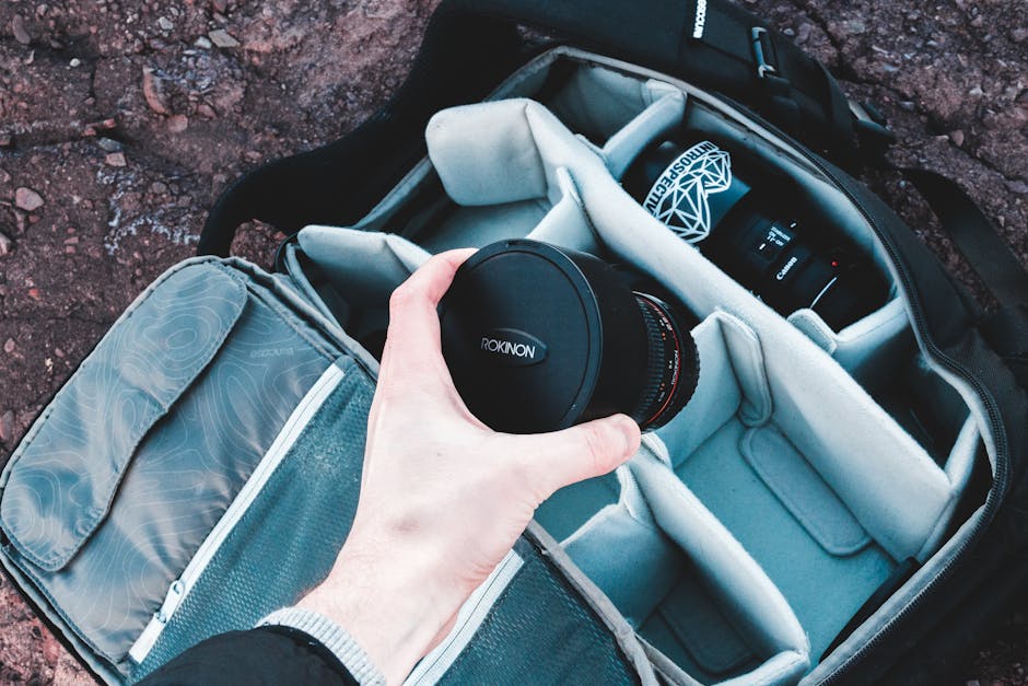 Hand holding Rokinon lens over an organized camera bag on rocky ground.
