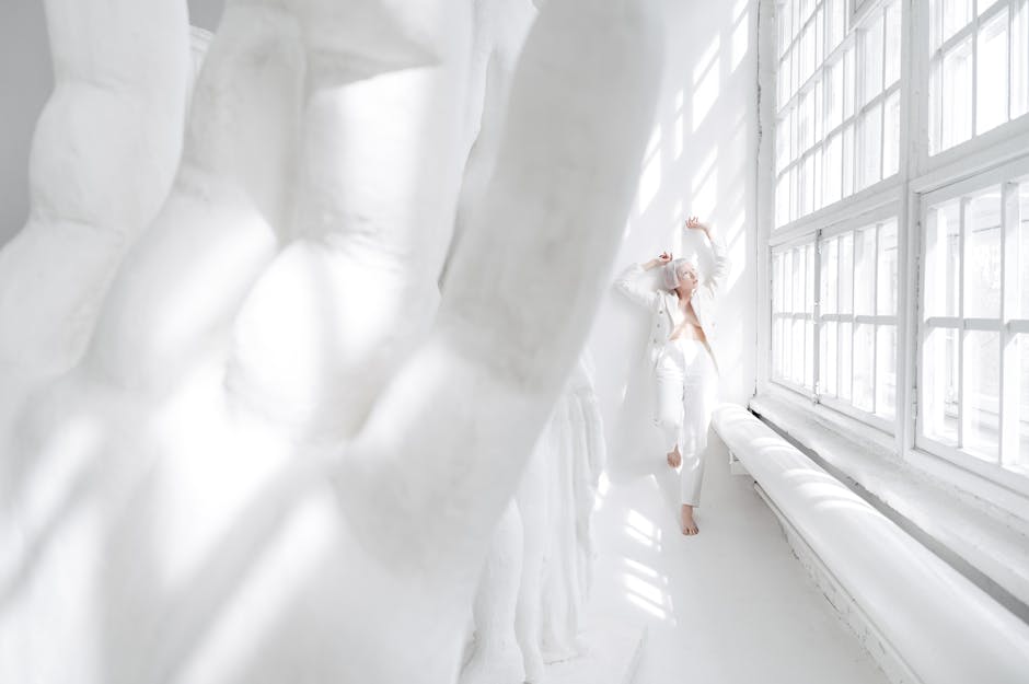 Minimalist fashion photo of a woman in white attire posing in a sunlit room with large windows.