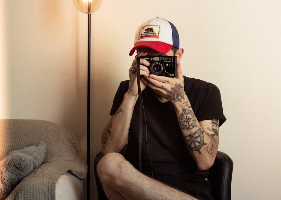 Tattooed photographer sitting indoors with camera, capturing a moment.