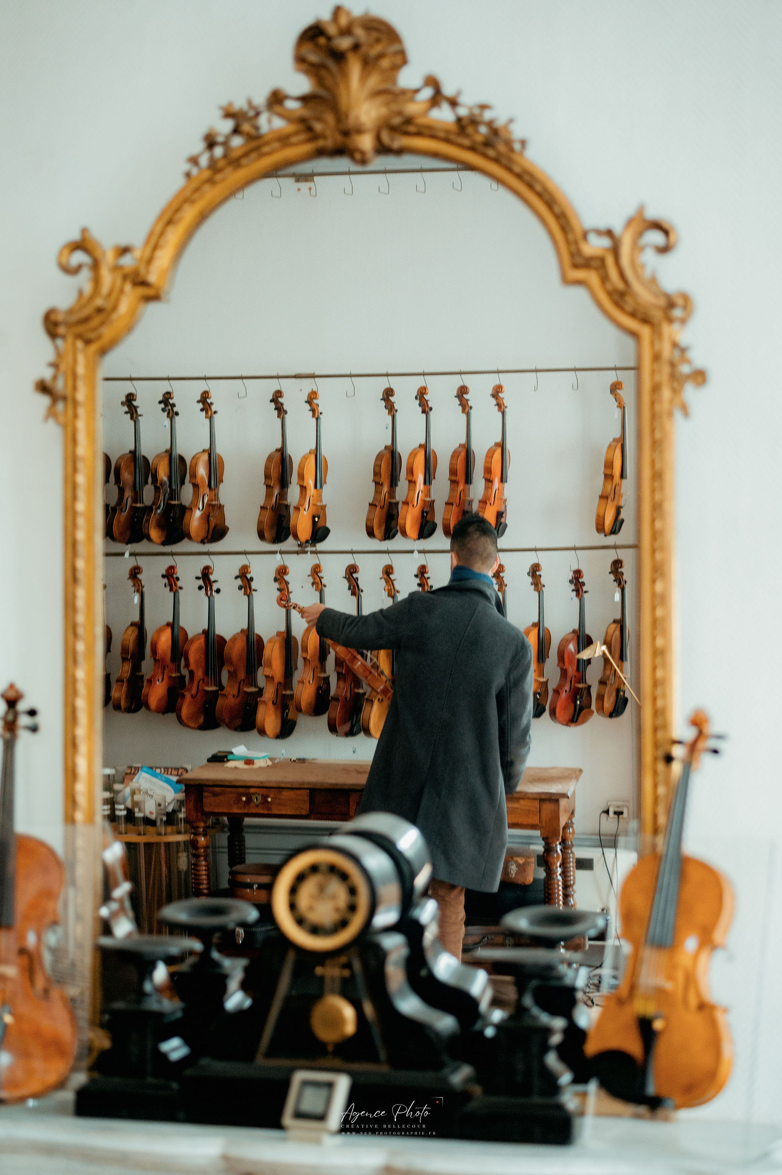 Luthier en veste noire sélectionnant un violon devant un mur de collection, encadré par un miroir doré orné