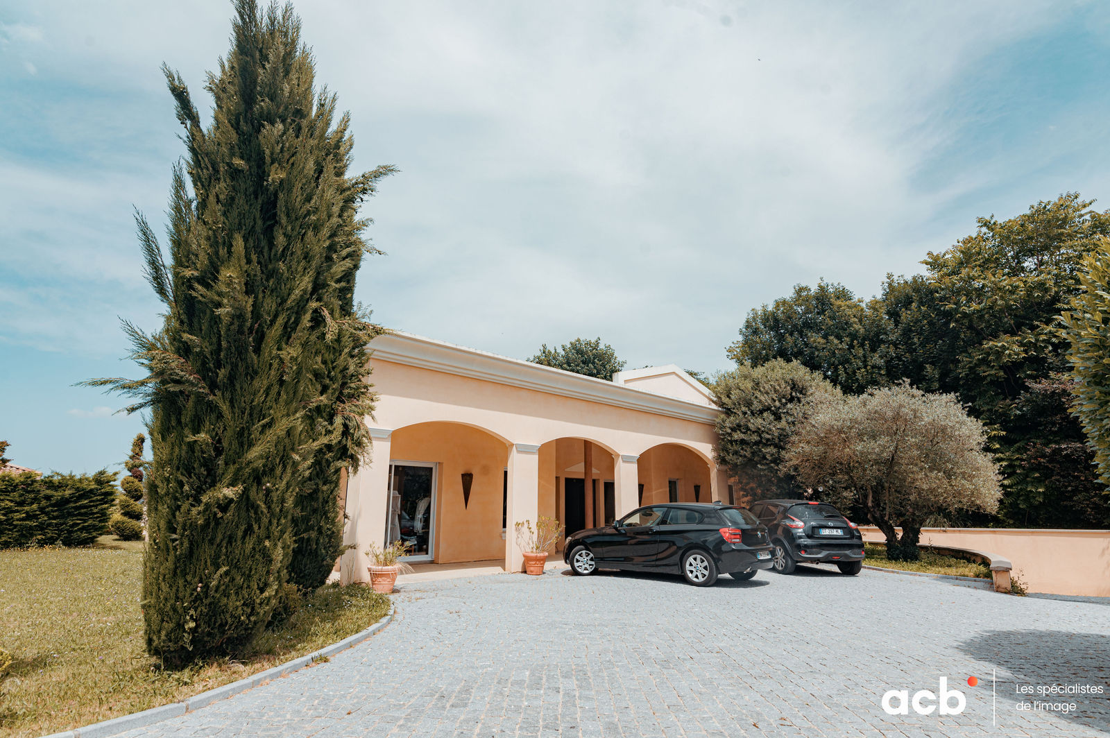 Villa blanche aux volets ocre avec cyprès et oliviers, allée pavée et deux voitures stationnées