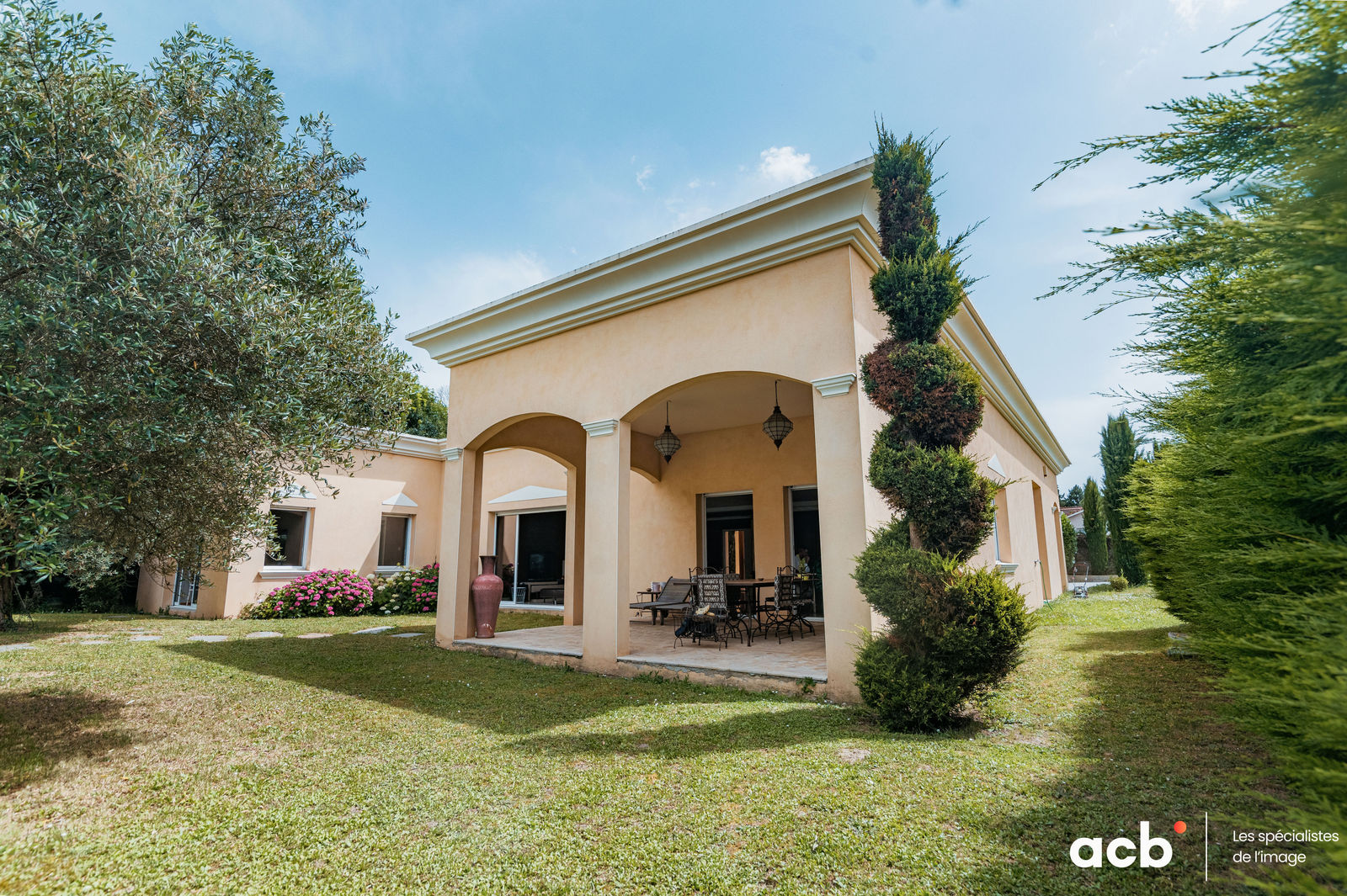 Maison beige avec porche voûté, terrasse ombragée et jardin paysager, cyprès et oliviers encadrant la façade