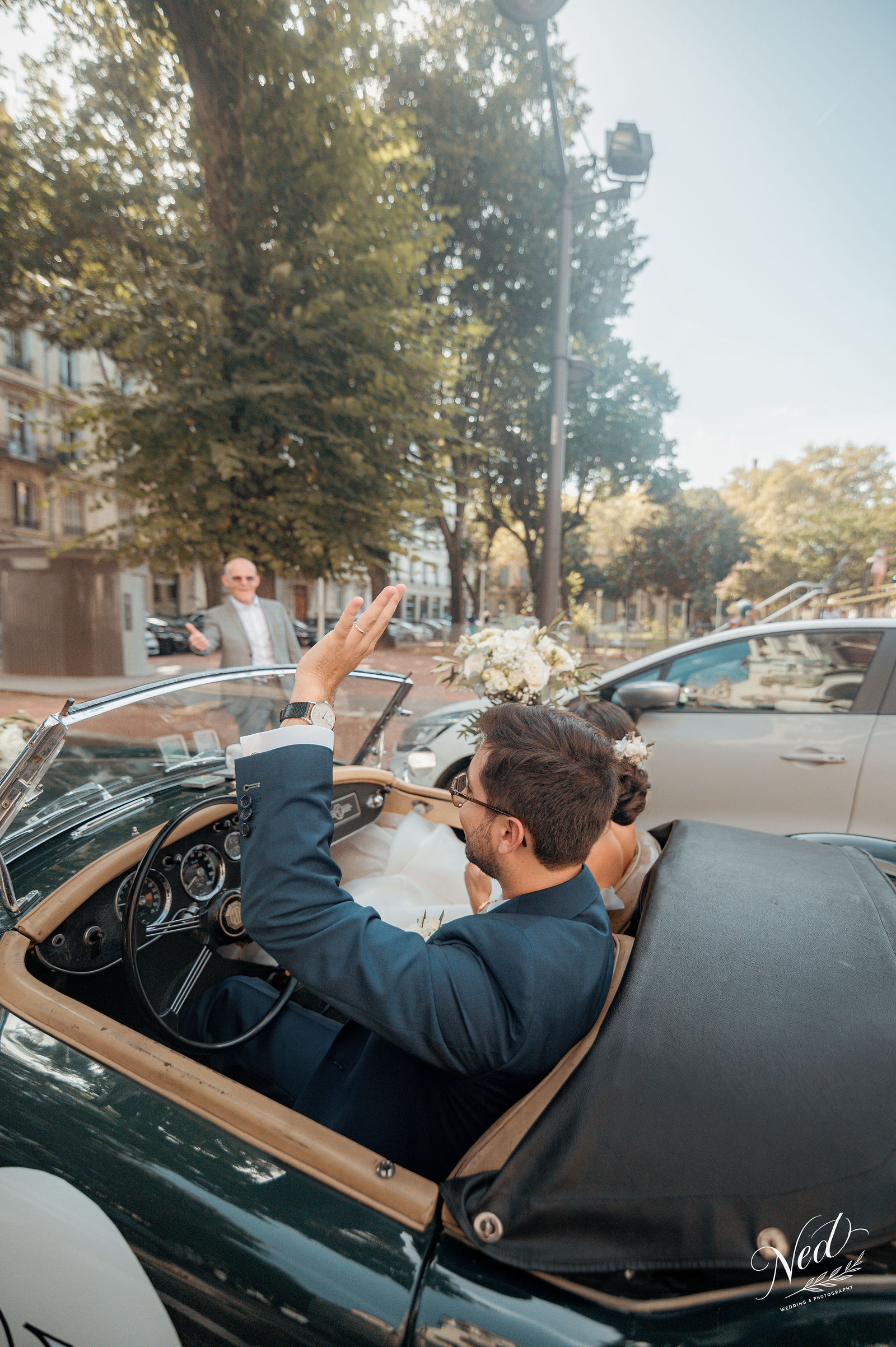 Mariés dans une décapotable vintage verte, le marié lève le bras en signe de joie sur une avenue borée d'arbres à Lyon