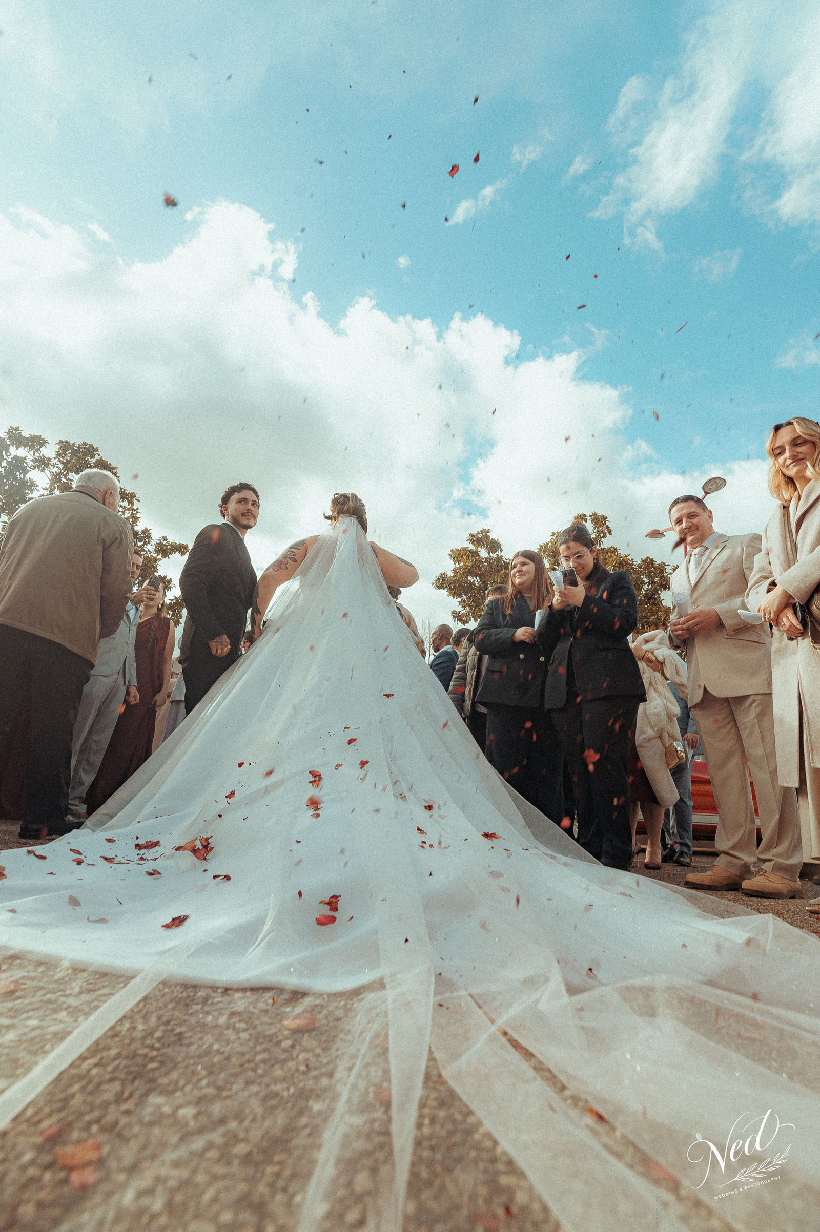 Mariée en robe blanche avec traîne longue, entourée de famille et amis qui lancent des pétales rouges, ciel bleu dégagé en arrière-plan
