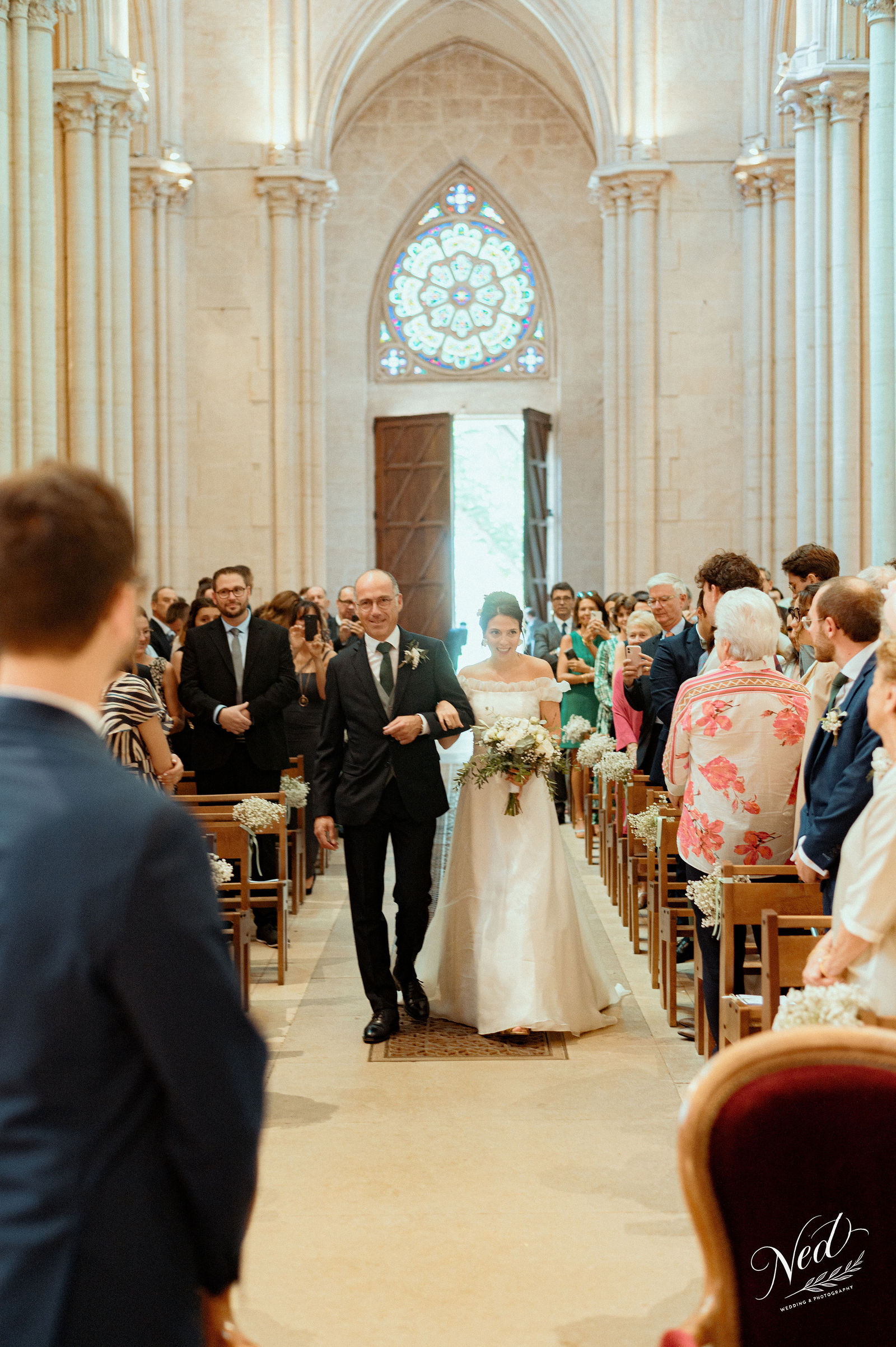 Mariée en robe blanche et marié en costume noir remontent l'allée d'une église gothique, invités debout de chaque côté applaudissant
