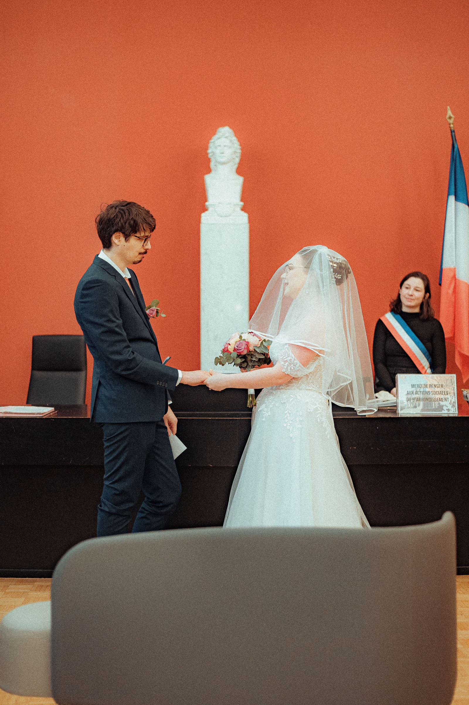 Mariée en robe blanche et voile reçoit son bouquet du marié en costume bleu à la mairie, maire en écharpe tricolore à droite