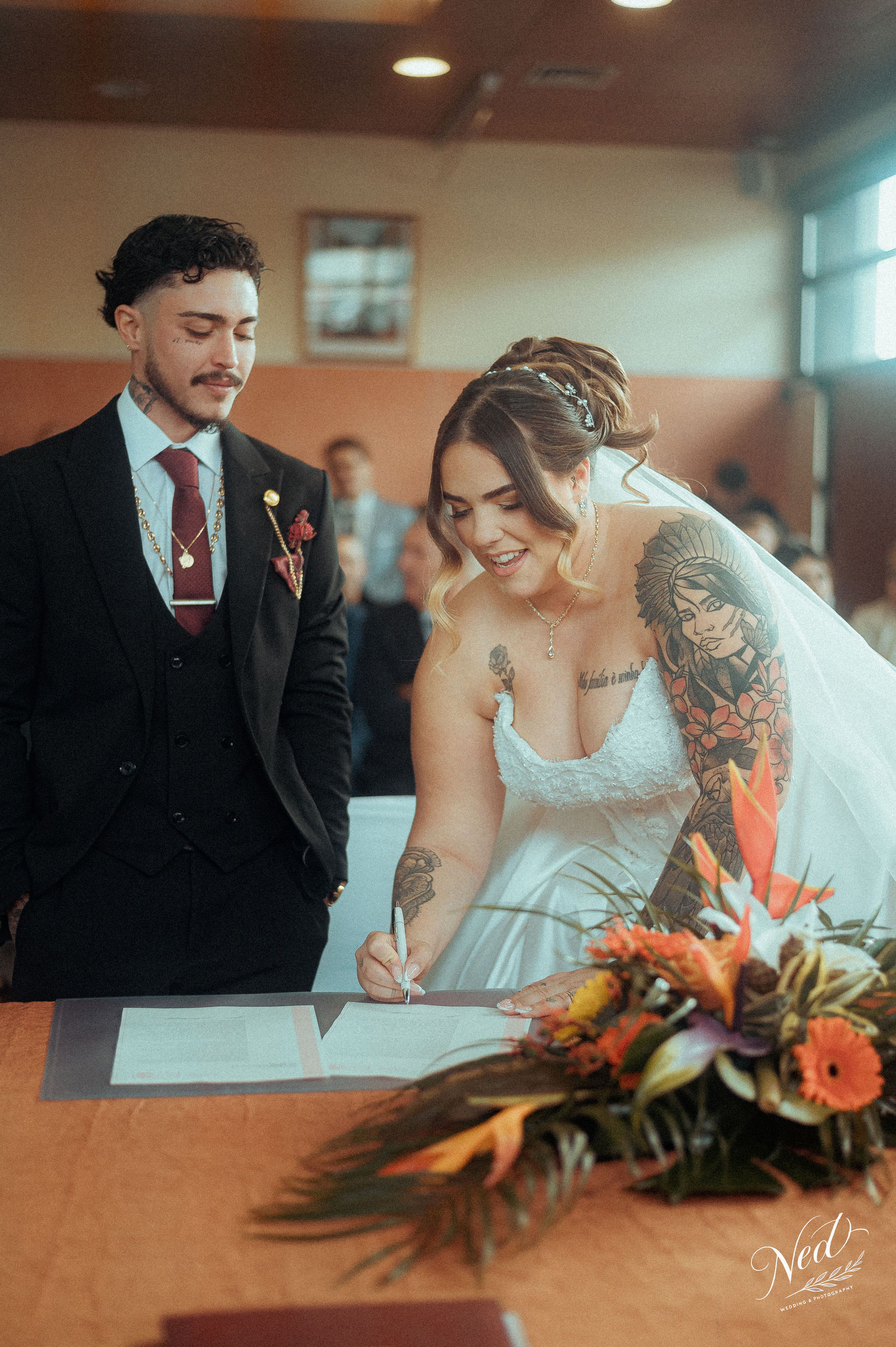 Mariée en robe blanche signe les registres à la mairie tandis que le marié en costume noir la regarde, bouquet de fleurs orange en premier plan