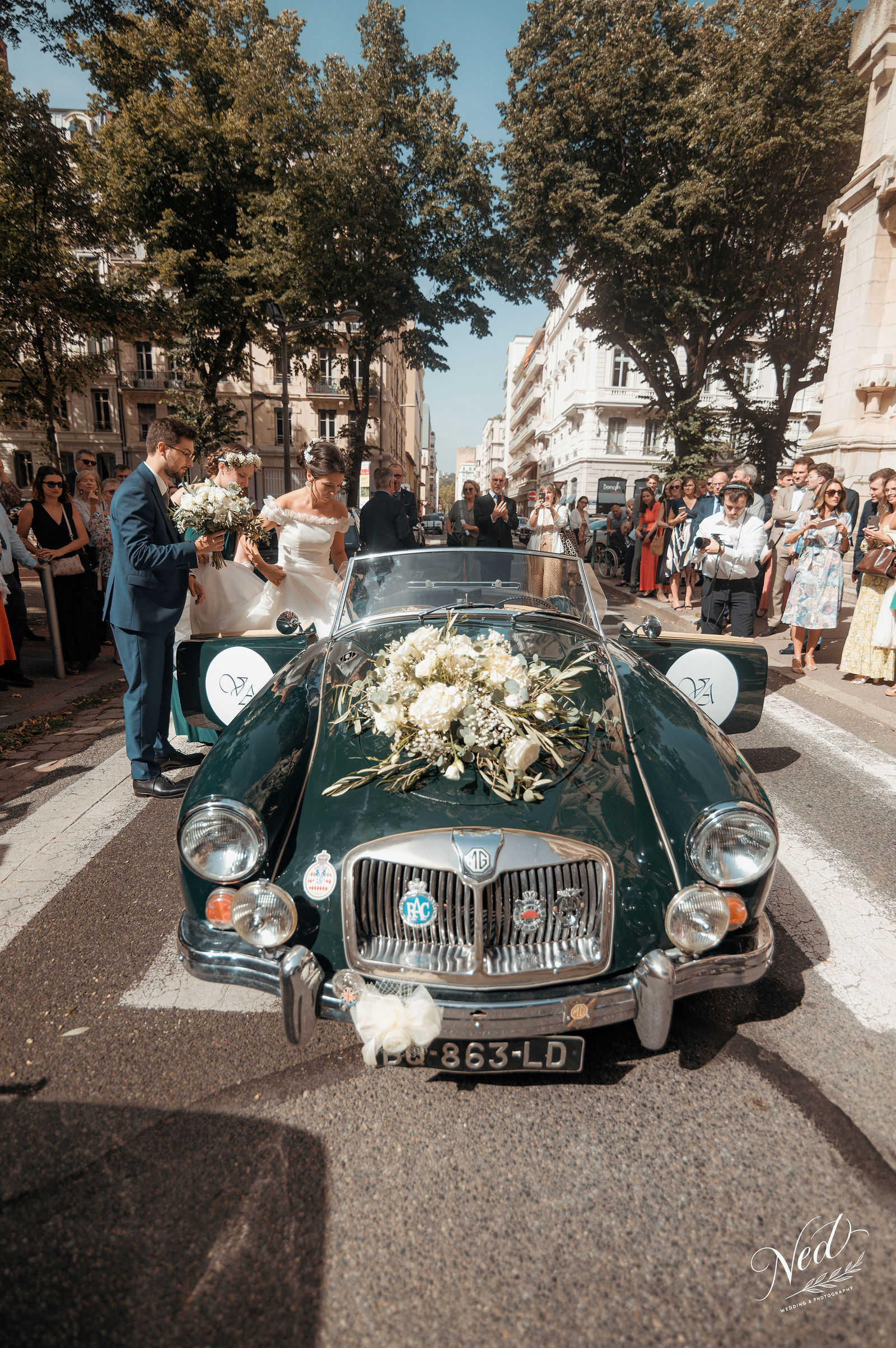 Couple de mariés dans une voiture décapotable verte vintage ornée de fleurs blanches, rue pavée bordée d'immeubles haussmanniens et d'invités souriants