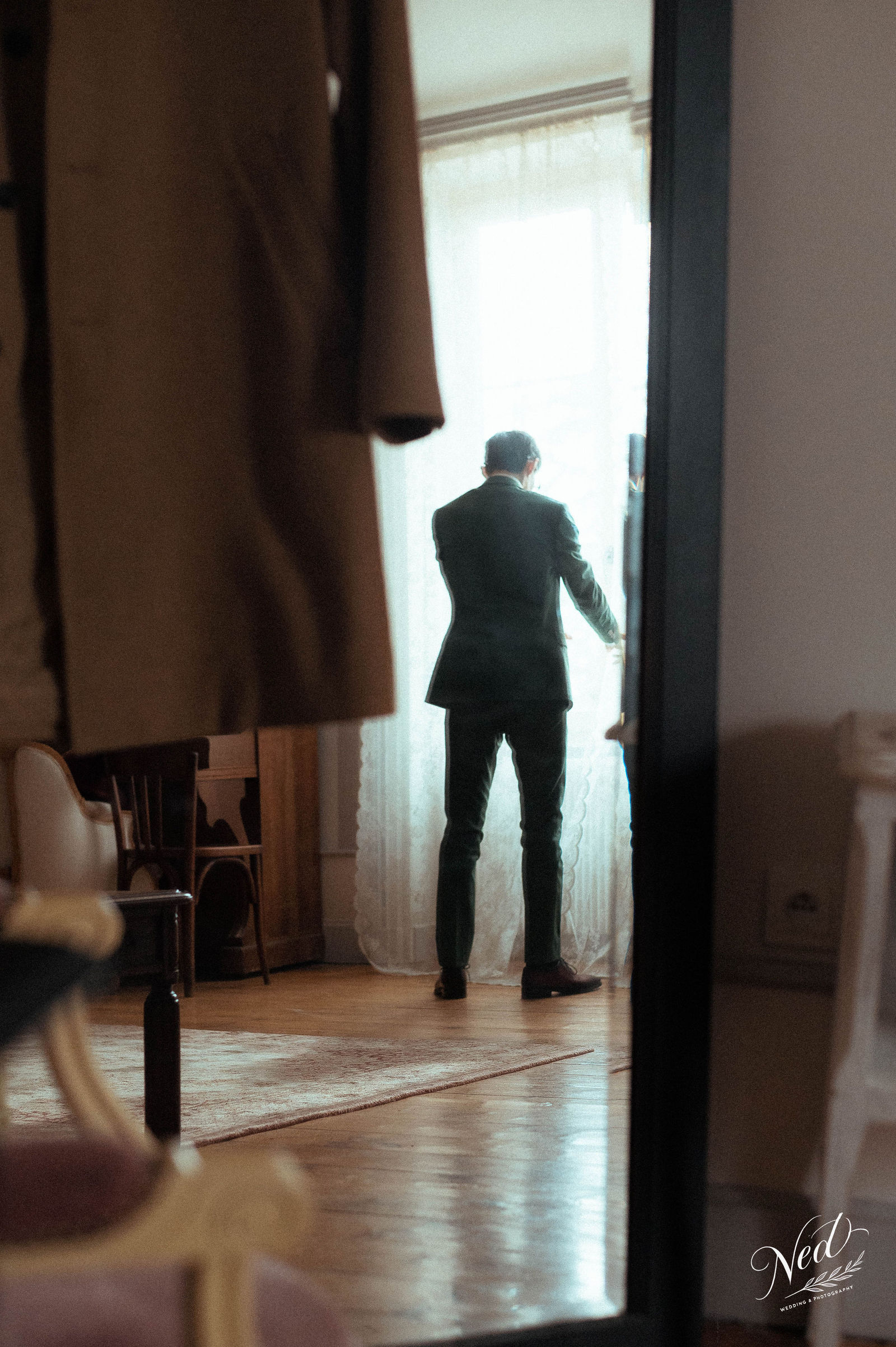 Homme en costume noir de dos regardant par la fenêtre d'une chambre, lumière naturelle intense