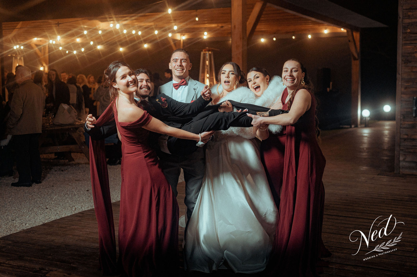 Mariée en robe blanche et marié en costume noir levés par six demoiselles d'honneur en robes bordeaux sous une structure en bois éclairée par des guirlandes lumineuses