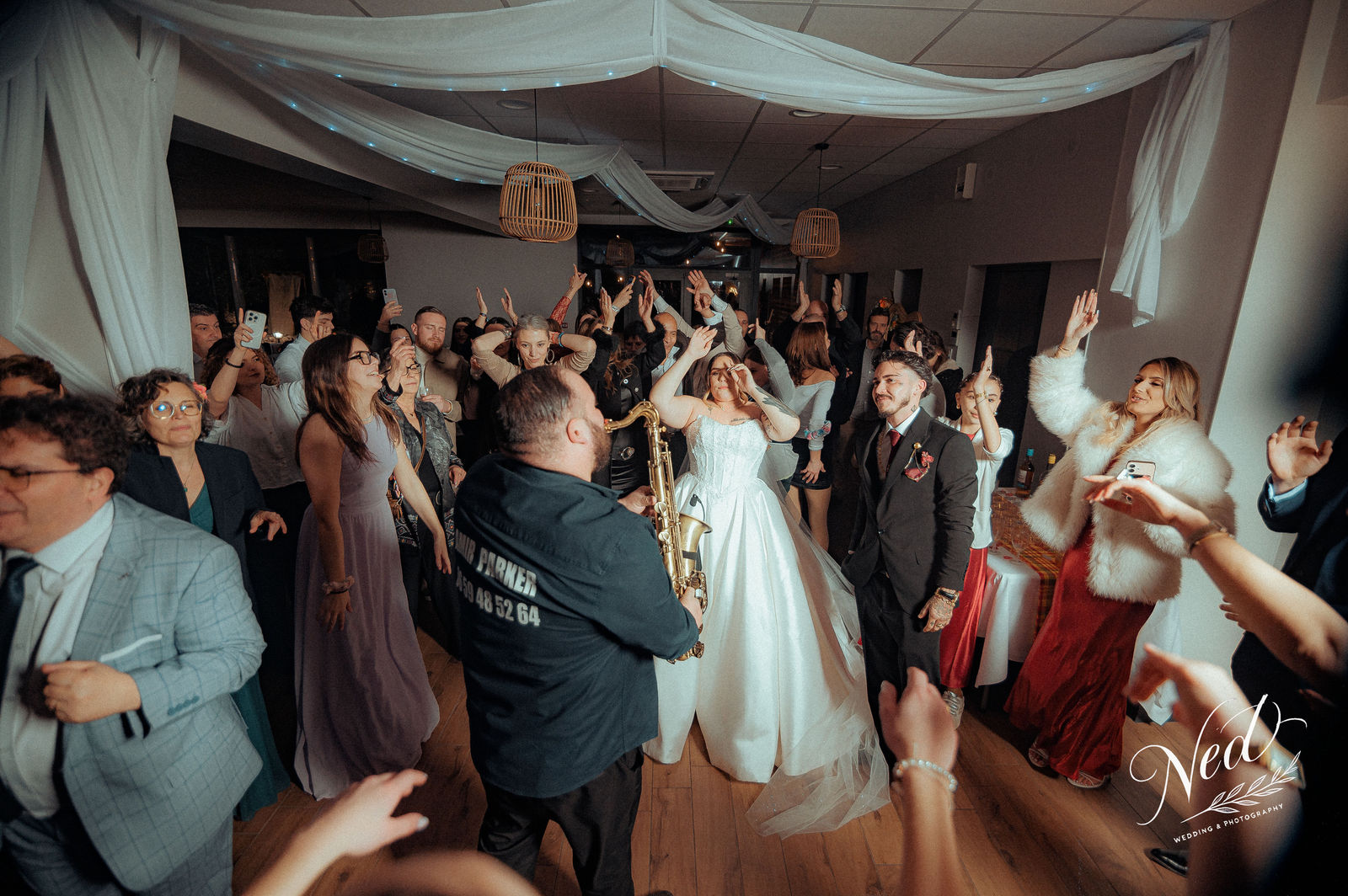 Mariée en robe blanche danse au milieu de la piste tandis qu'un saxophoniste joue, les invités lèvent les bras autour d'eux