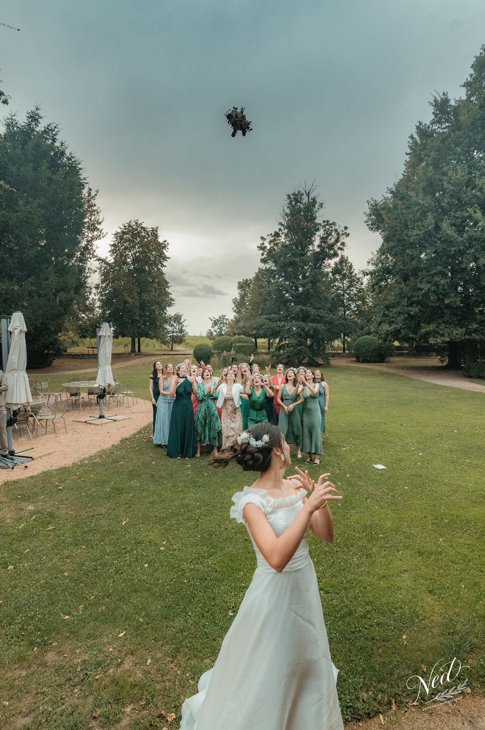 Mariée en robe blanche lance son bouquet vers un groupe de demoiselles d'honneur en robes vertes, dans un parc arboré