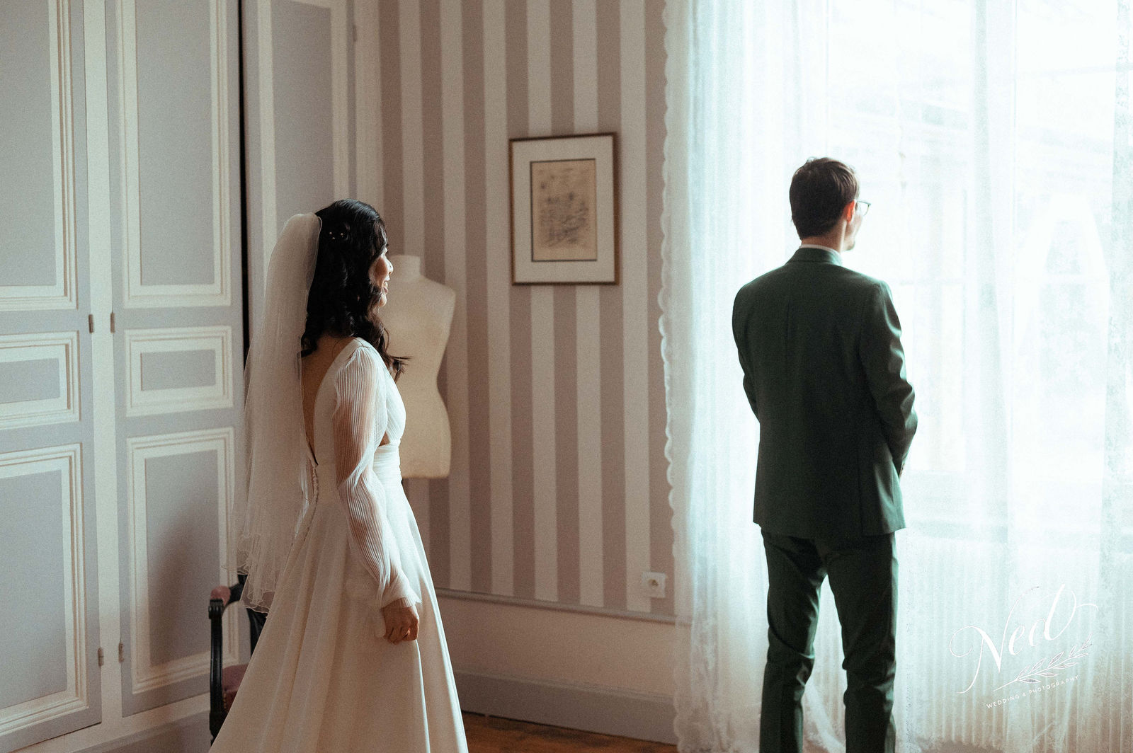 Mariée en robe blanche et voile face à son futur mari en costume vert, dos à dos dans un salon élégant avec fenêtres lumineuses