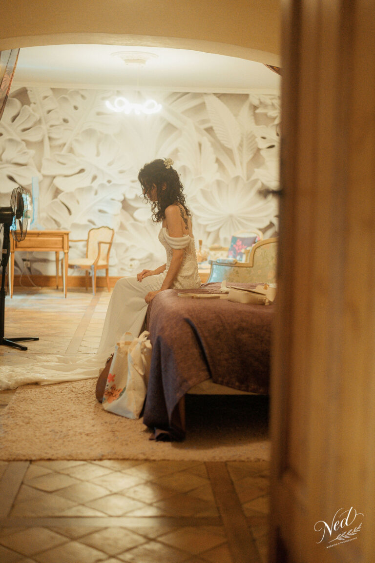 Mariée en robe blanche assise sur un fauteuil dans une salle décorée, photographiée à travers une porte ouverte