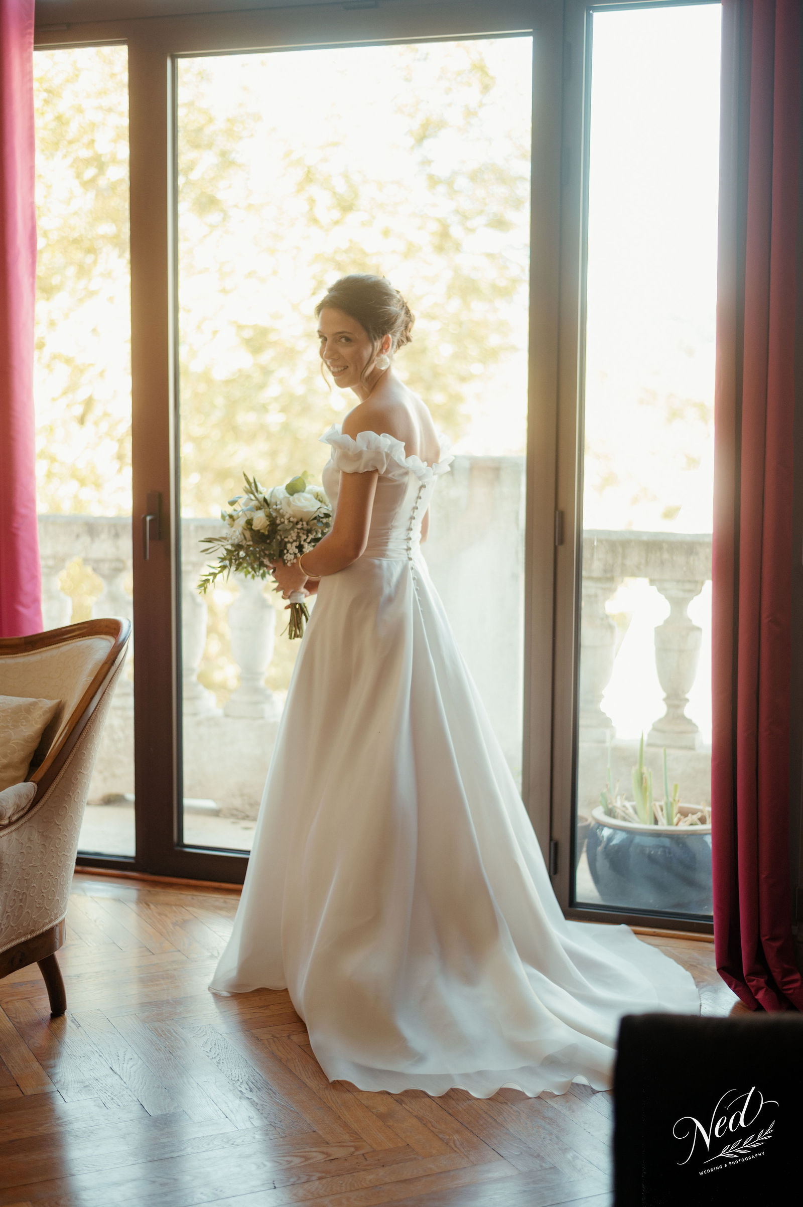 Mariée en robe blanche avec bouquet, souriante de profil devant une baie vitrée avec vue sur la nature