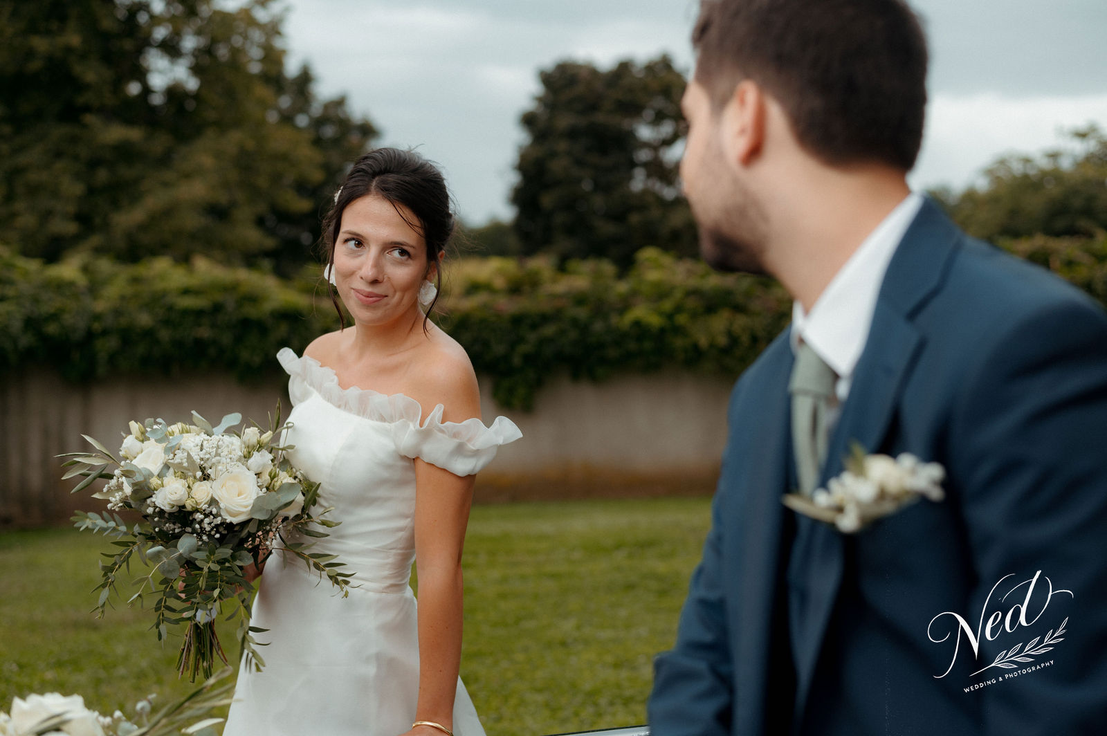 Mariée en robe blanche avec bouquet de roses regarde son mari en costume bleu marine dans un jardin