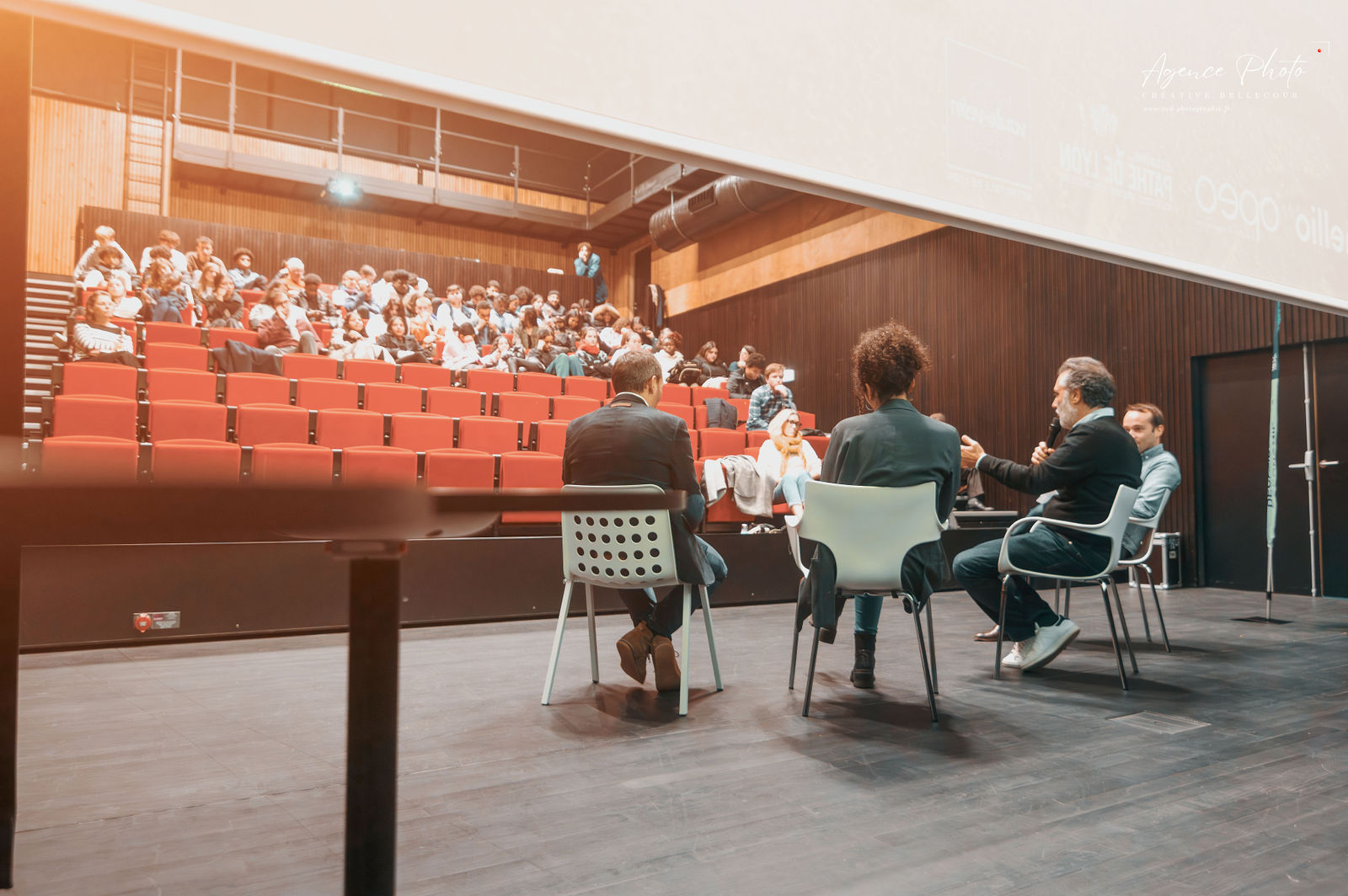 Trois panélistes assis face à une audience de cinquante personnes dans un amphithéâtre aux sièges rouges