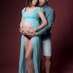 Femme enceinte en robe bleu ciel et son partenaire enlacés de face sur fond bordeaux