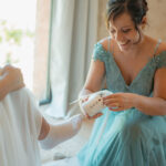 Femme enceinte en robe bleu ciel sourit en tenant un body blanc, lumière naturelle du jour