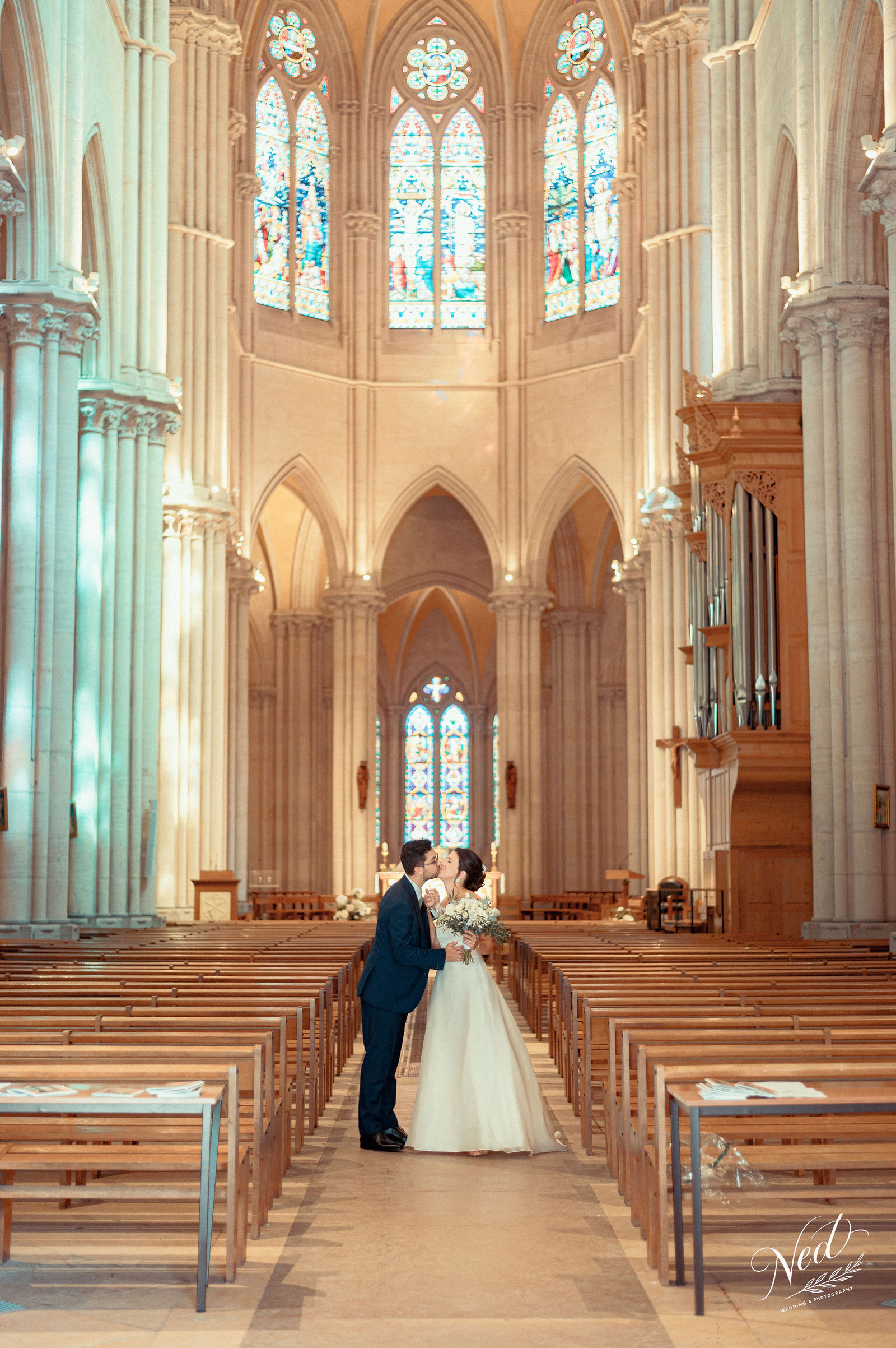 Couple de mariés s'embrassant dans la nef d'une cathédrale gothique, vitraux bleus en arrière-plan, lumière naturelle dorée
