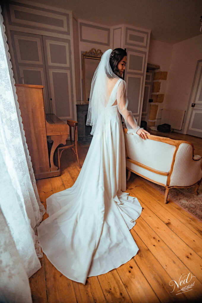 Jeune mariée de profil en robe blanche fluide et voile, posant dans un salon classique aux portes ornementales, parquet bois clair