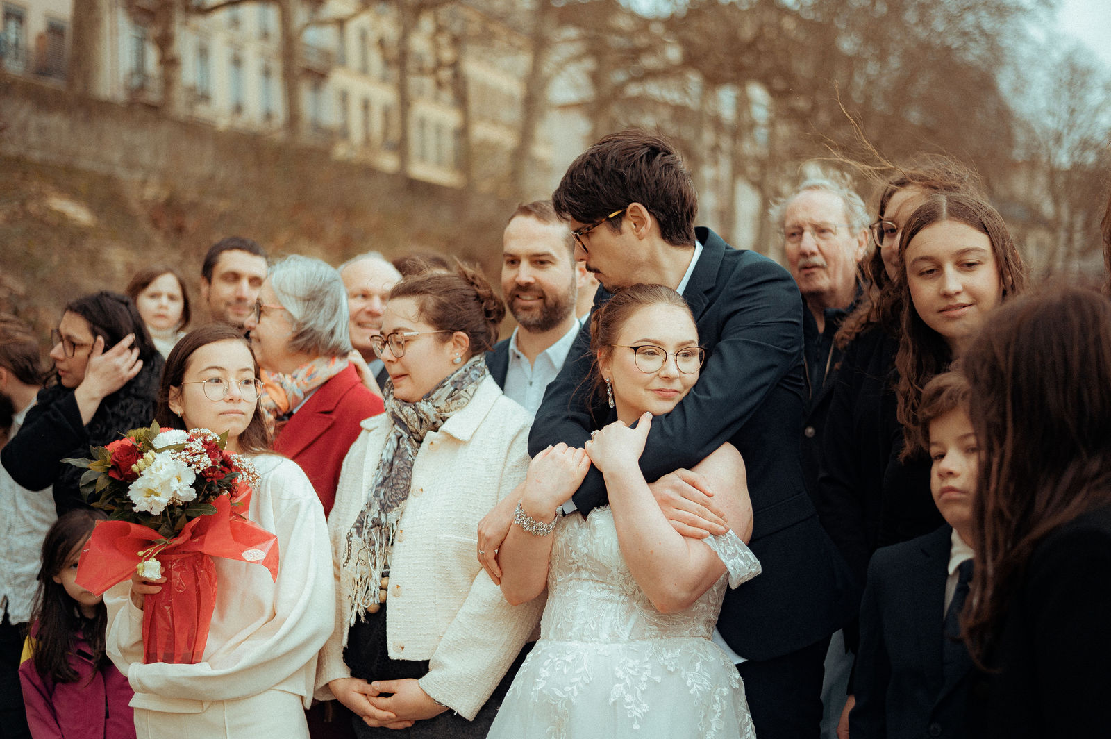 Mariée en robe blanche avec bouquet rouge entourée de sa famille et amis devant des immeubles lyonnais en hiver