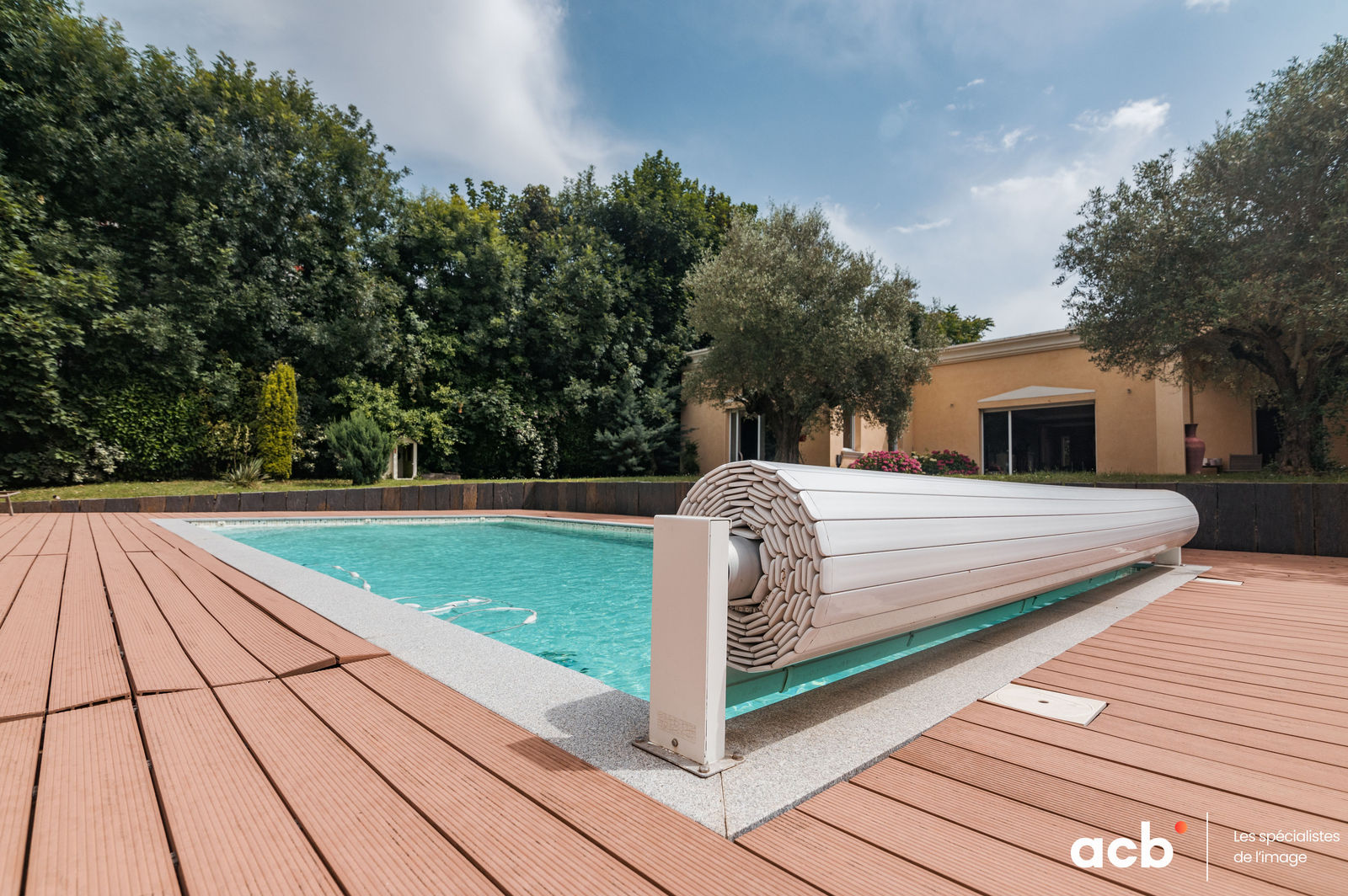 Piscine aux eaux turquoise avec volet roulant blanc, terrasse en bois composite, maison ocre et arbres verts en arrière-plan