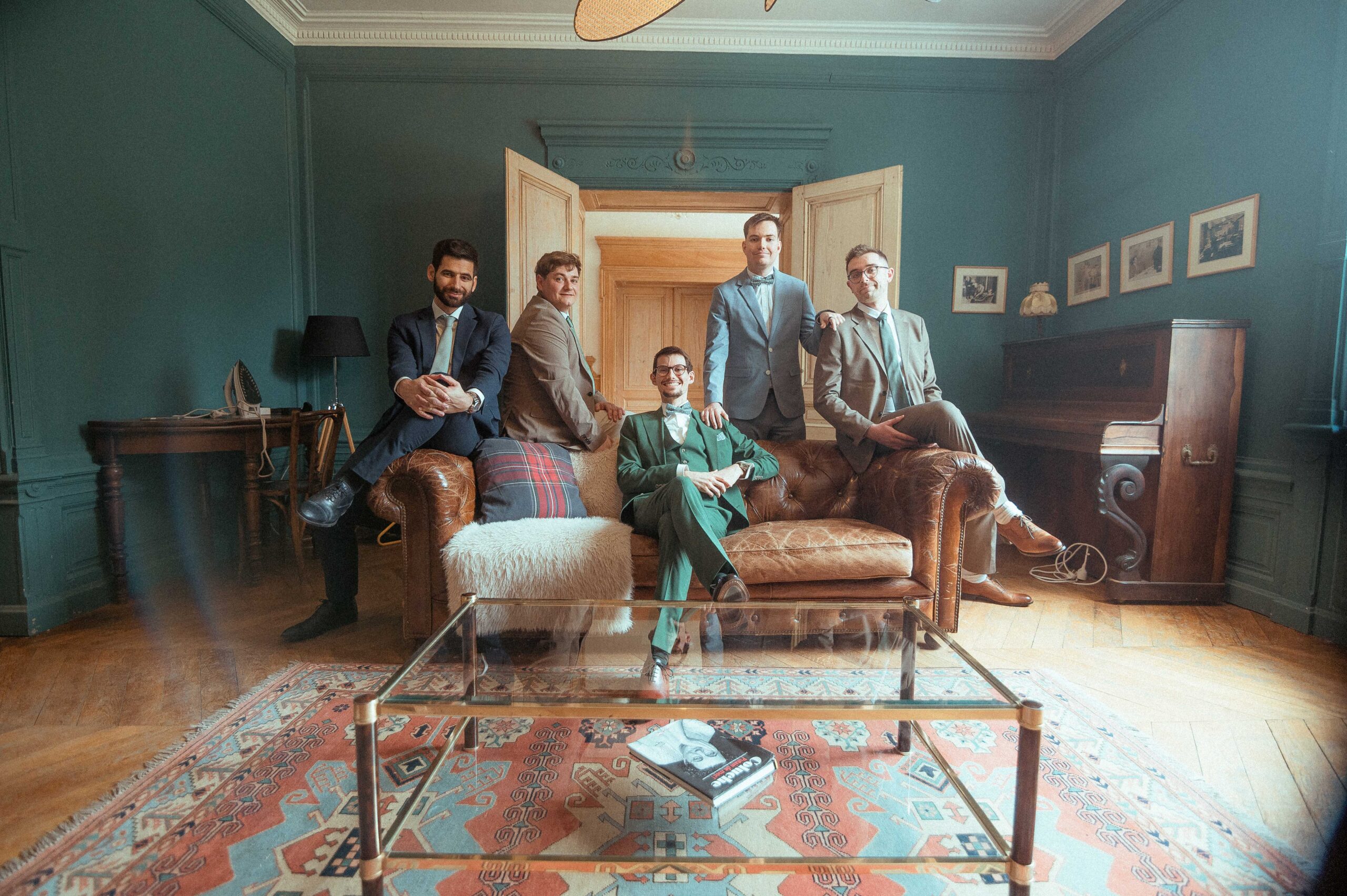 Cinq hommes en costumes posent dans un salon aux murs bleu-vert, assis et debout autour de fauteuils en cuir marron