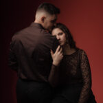 Homme en chemise noire embrassant une femme en dentelle, fond rouge bordeaux, portrait studio