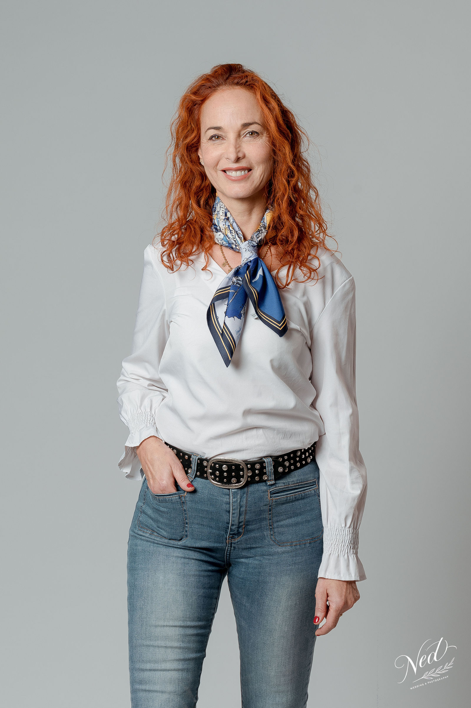 Femme aux cheveux roux ondulés sourit face à l'appareil photo, chemise blanche et foulard bleu noué au cou.