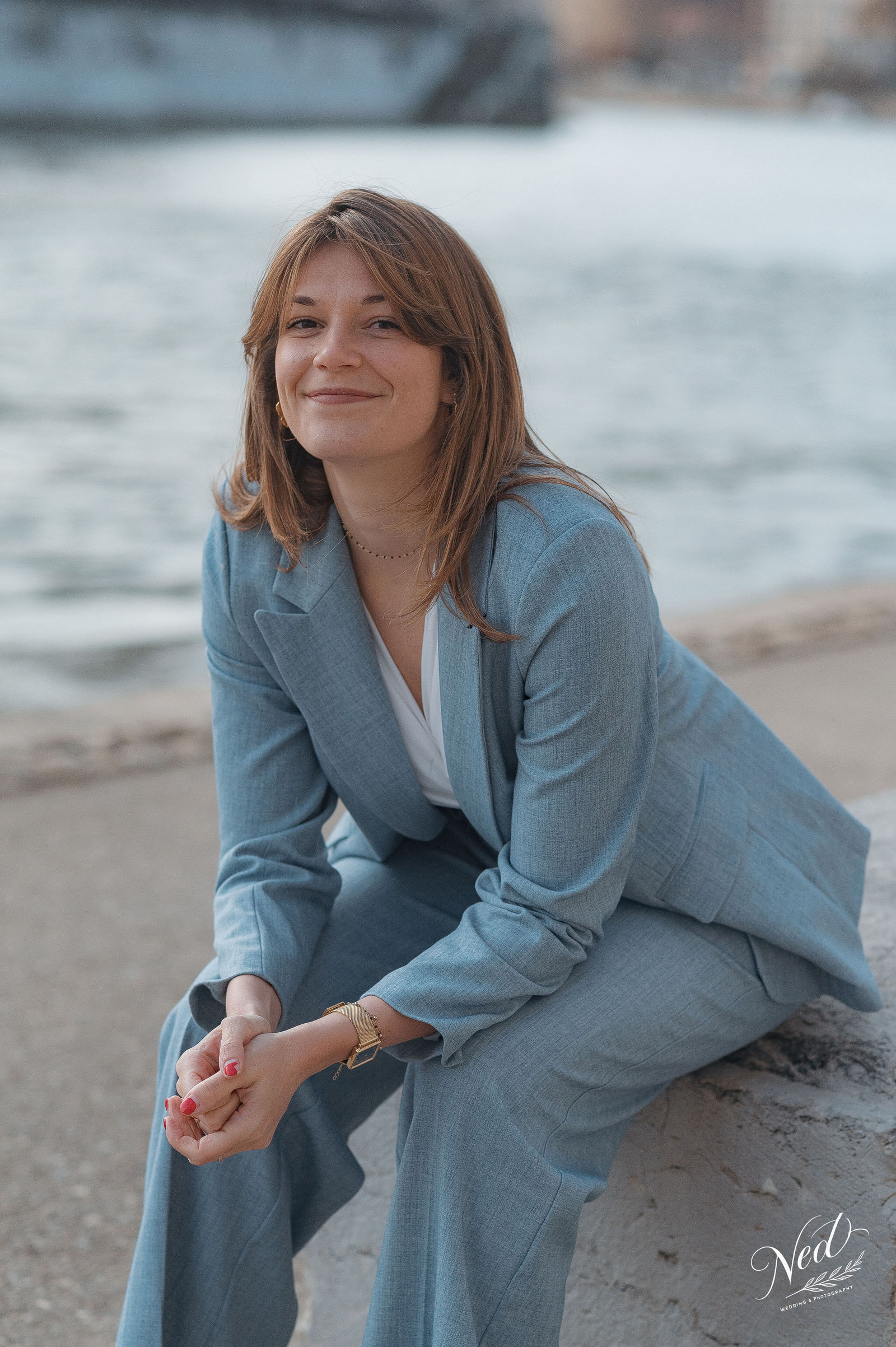 Femme en costume bleu ciel posée sur la plage, eau floue en arrière-plan, lumière naturelle douce