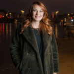 Jeune femme aux cheveux roux sourit face à la caméra, vêtue d'un manteau kaki, les lumières de la ville floutées en arrière-plan la nuit