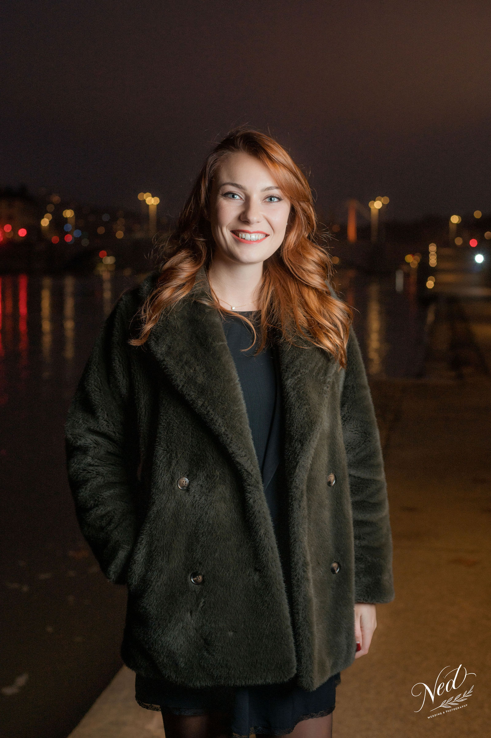 Jeune femme aux cheveux roux sourit face à la caméra, vêtue d'un manteau kaki, les lumières de la ville floutées en arrière-plan la nuit