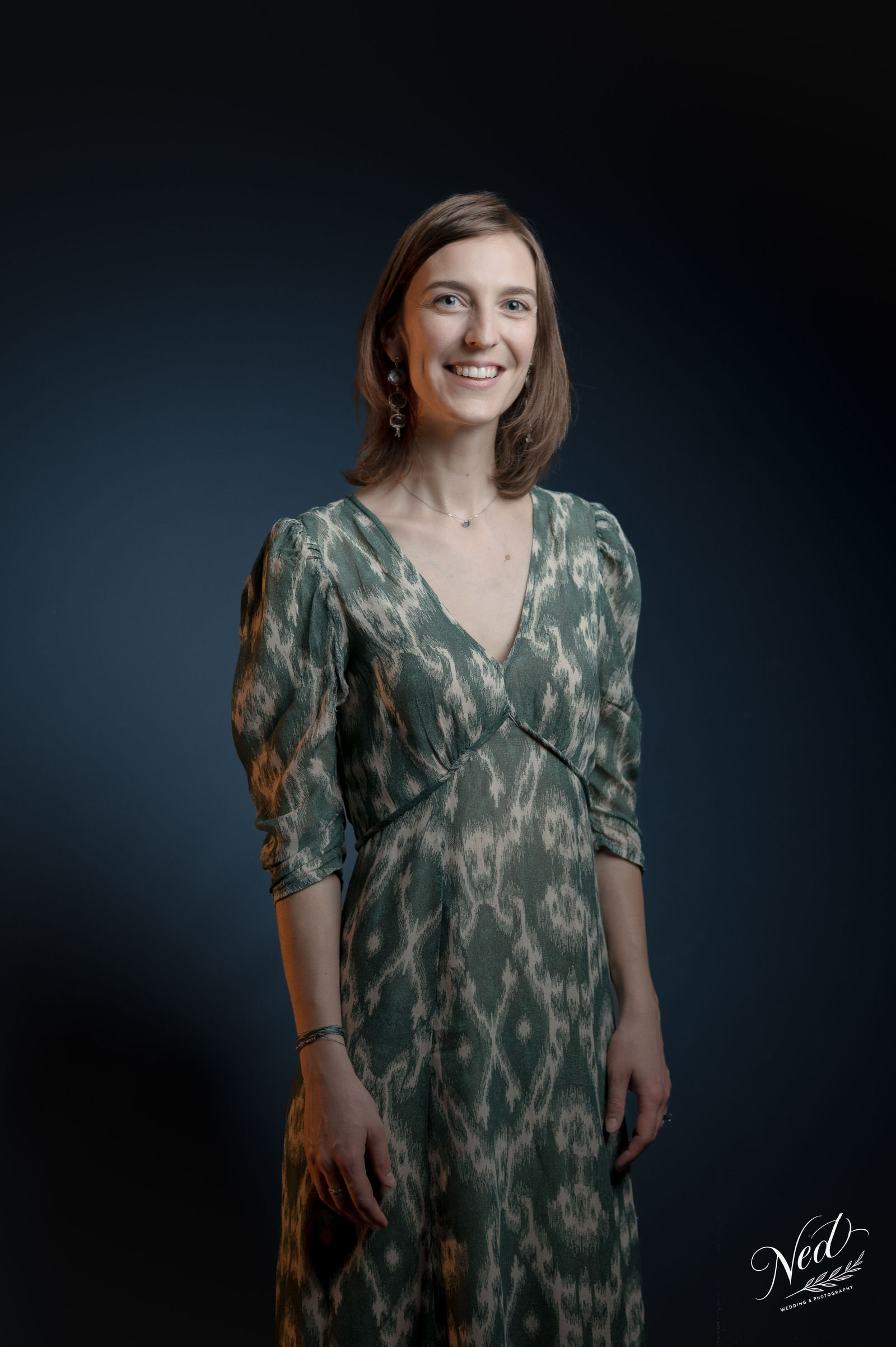 Jeune femme souriante en robe verte motifs géométriques, debout face à la caméra sur fond bleu foncé