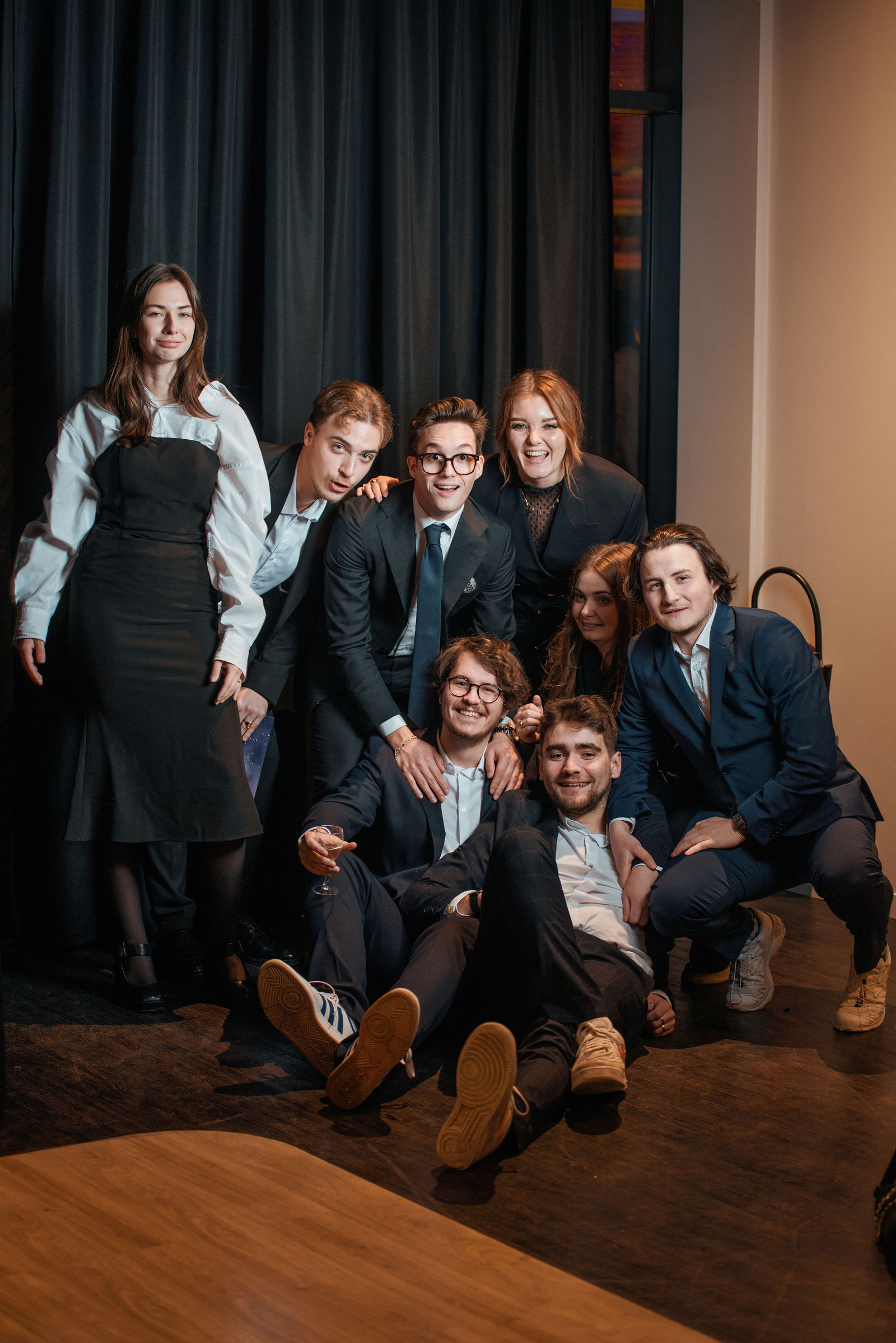 Sept jeunes adultes en costumes noirs et blancs posent ensemble dans un studio, certains assis d'autres debout, souriant à la caméra