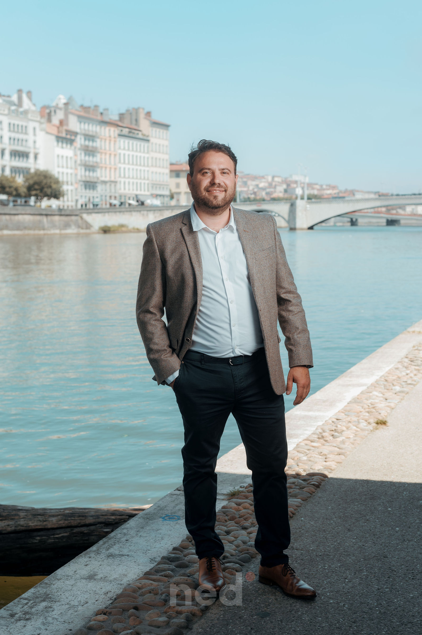 Homme en blazer beige et chemise blanche posant sur les berges du Rhône à Lyon, pont et immeubles en arrière-plan