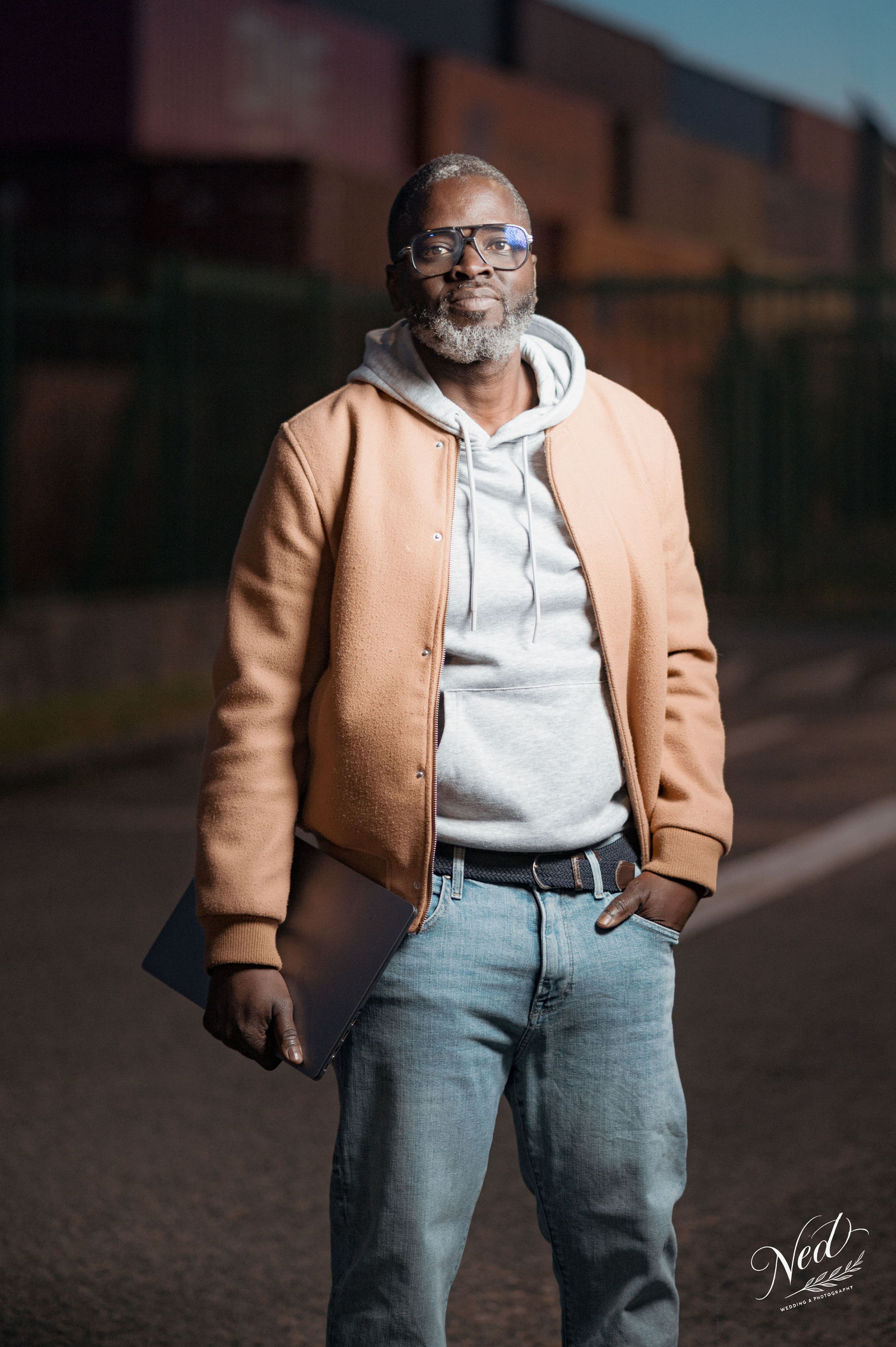 Homme portant veste camel et hoodie gris pose devant bâtiment en brique, lumière naturelle latérale