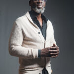 Homme portant des lunettes et un cardigan crème sur chemise noire, pantalon marron clair, pose de trois quarts sur fond gris
