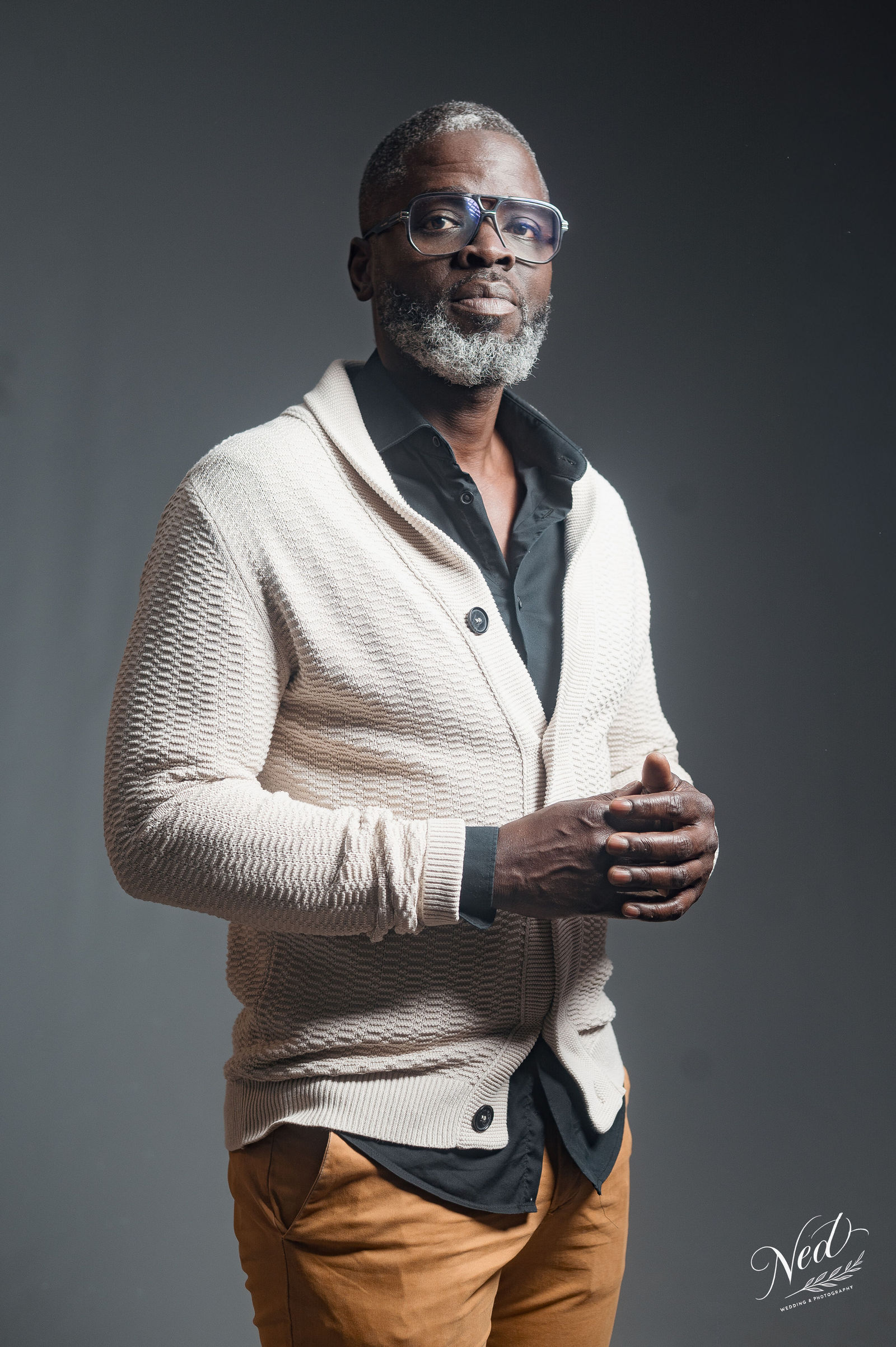 Homme portant des lunettes et un cardigan crème sur chemise noire, pantalon marron clair, pose de trois quarts sur fond gris