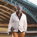 Homme en cardigan blanc et pantalon camel pose sous un pont en pierre à Lyon, lunettes teintées, sac à la main