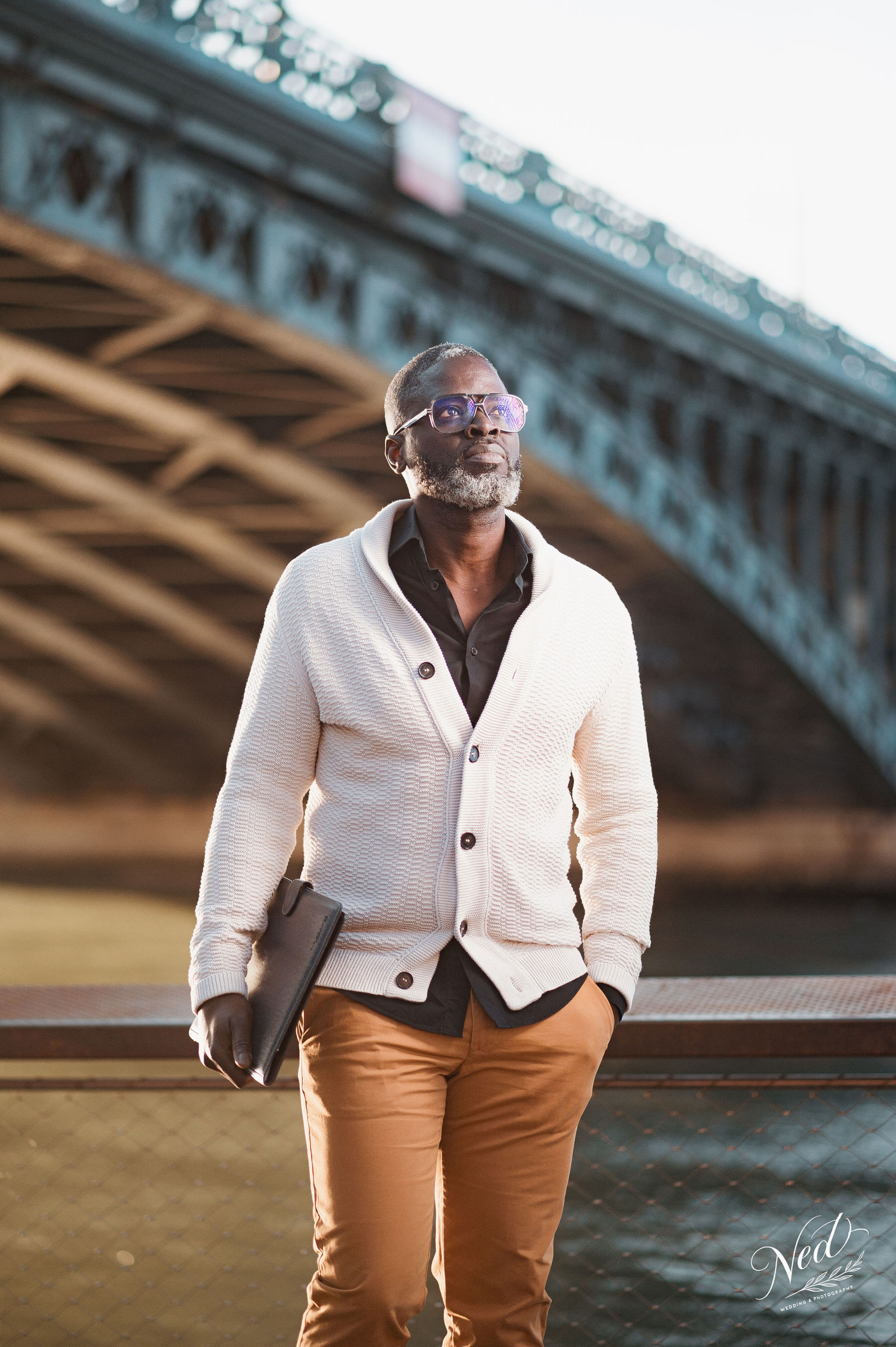 Homme en cardigan blanc et pantalon camel pose sous un pont en pierre à Lyon, lunettes teintées, sac à la main