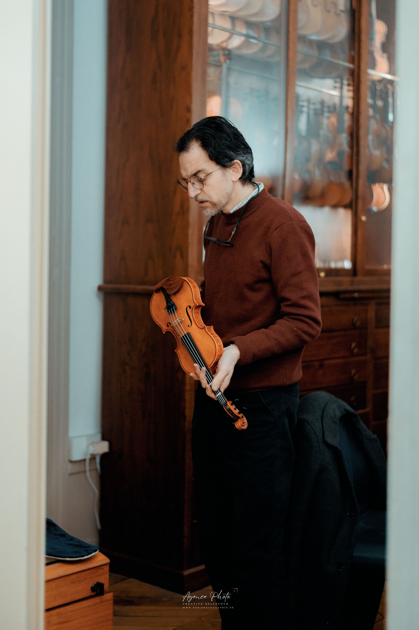 Homme en pull marron tenant un violon, regardant vers le bas, debout devant une porte en bois dans un intérieur.