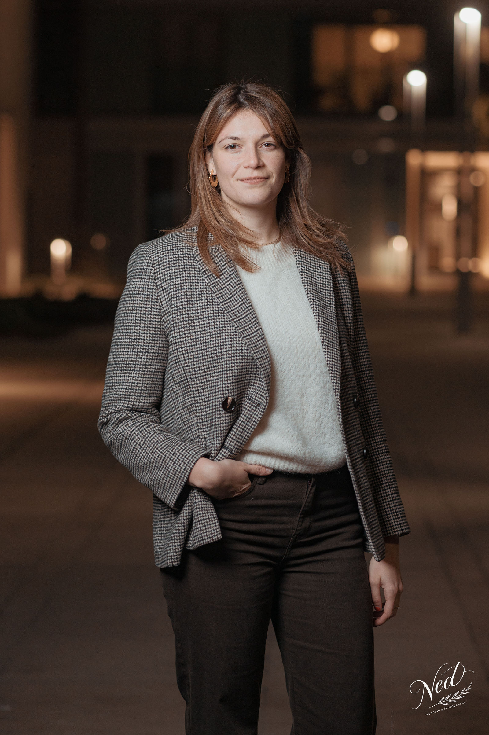 Femme en blazer gris à carreaux et pull blanc pose de nuit en extérieur, lumière urbaine douce en arrière-plan