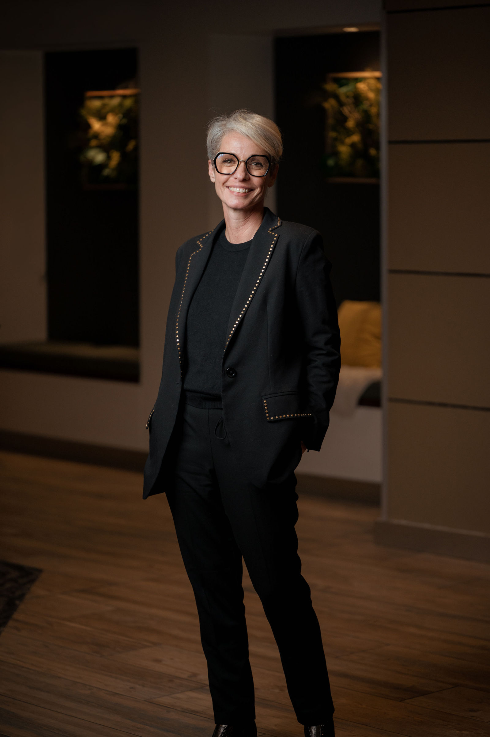 Femme aux cheveux courts gris sourit face à la caméra, vêtue d'un costume noir avec détails cloutés dorés, intérieur moderne
