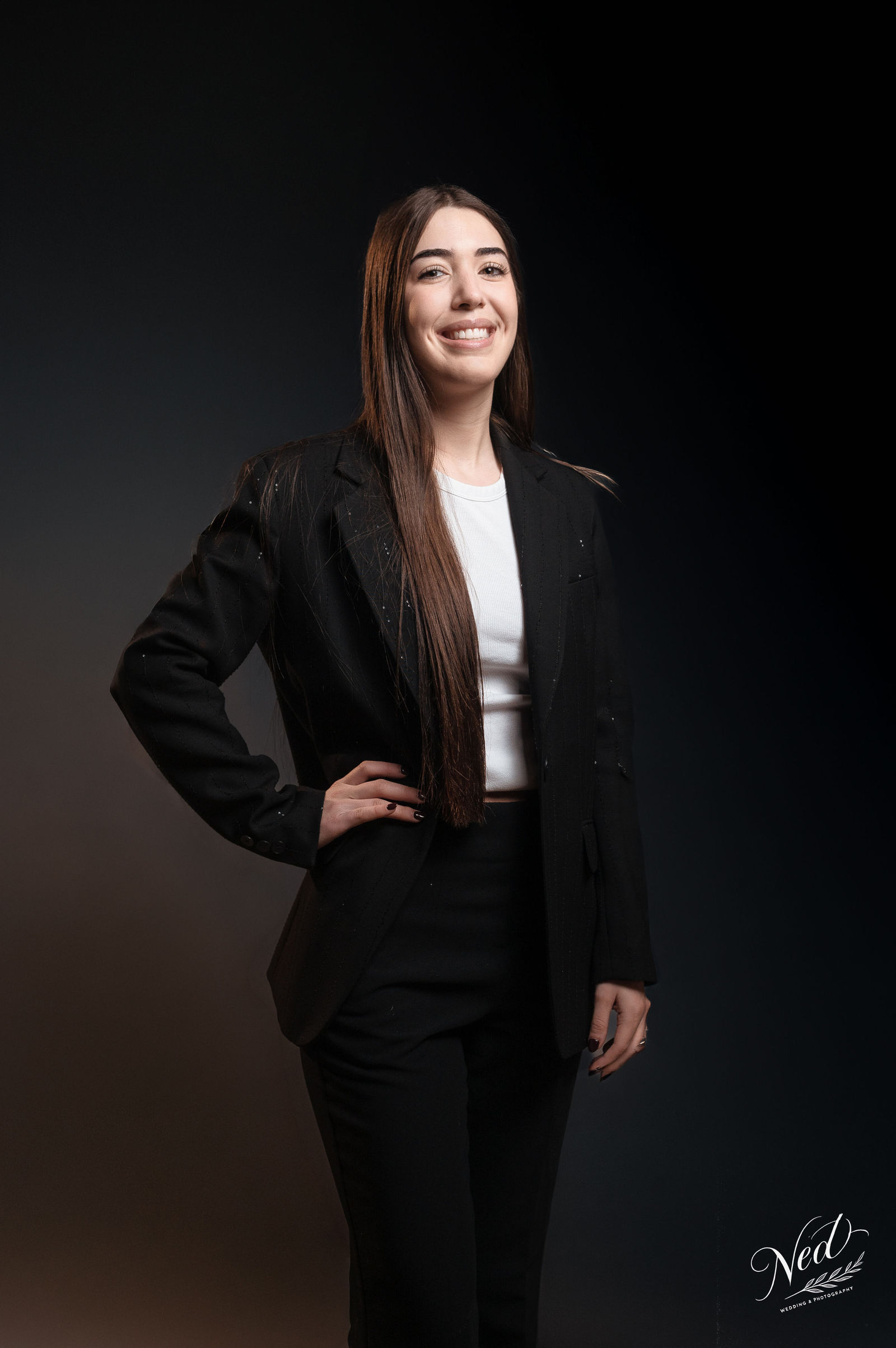 Femme souriante en blazer noir et top blanc, posant de profil sur fond noir uni
