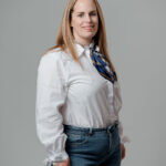 Femme aux cheveux roux souriante, chemise blanche et foulard bleu, posture de trois quarts face à la caméra