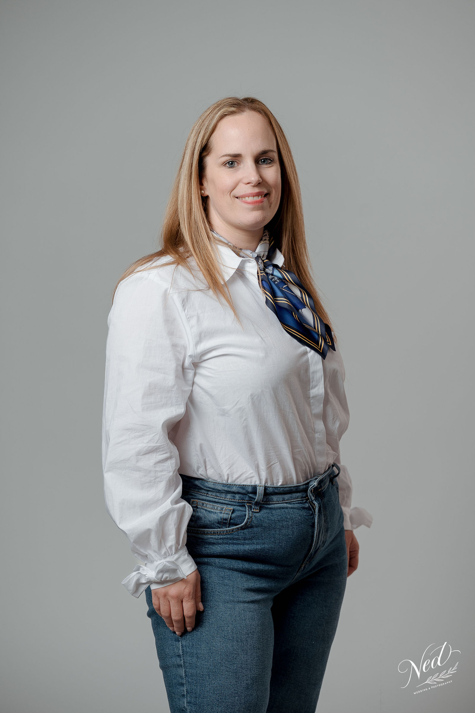 Femme aux cheveux roux souriante, chemise blanche et foulard bleu, posture de trois quarts face à la caméra