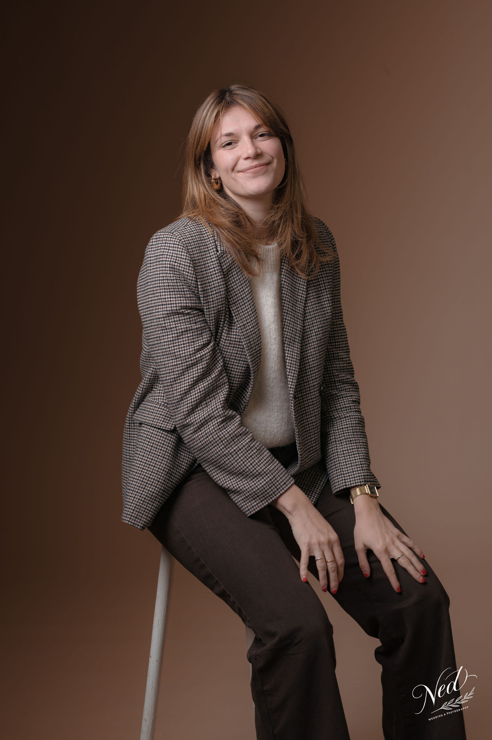 Femme souriante assise sur une chaise, veste à carreaux grise et pantalon noir, fond marron clair